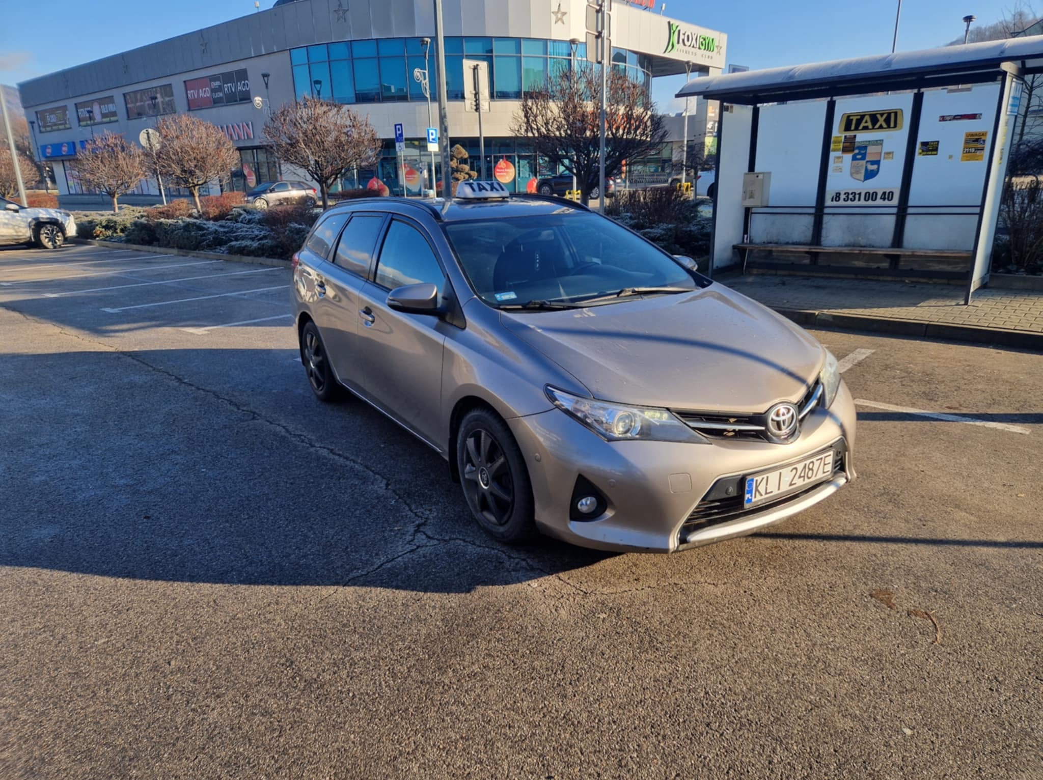 Szary samochód kombi z napisem TAXI na dachu, zaparkowany na parkingu przed budynkiem z szyldem 'FOXSIM FITNESS CLUB', obok przystanku autobusowego TAXI.