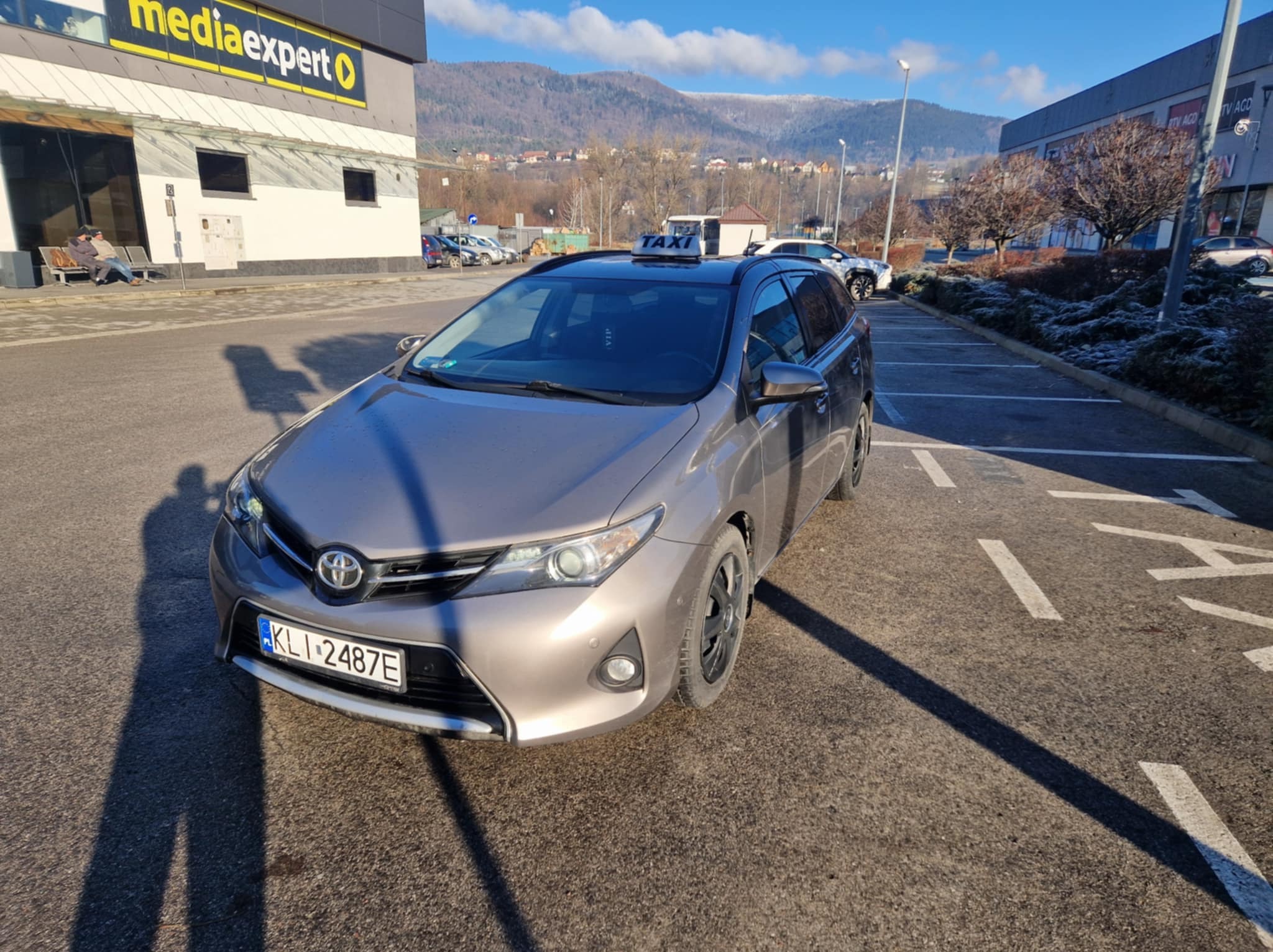Szary samochód typu kombi z napisem TAXI na dachu, zaparkowany na parkingu przed budynkiem Media Expert, w tle widoczne góry.
