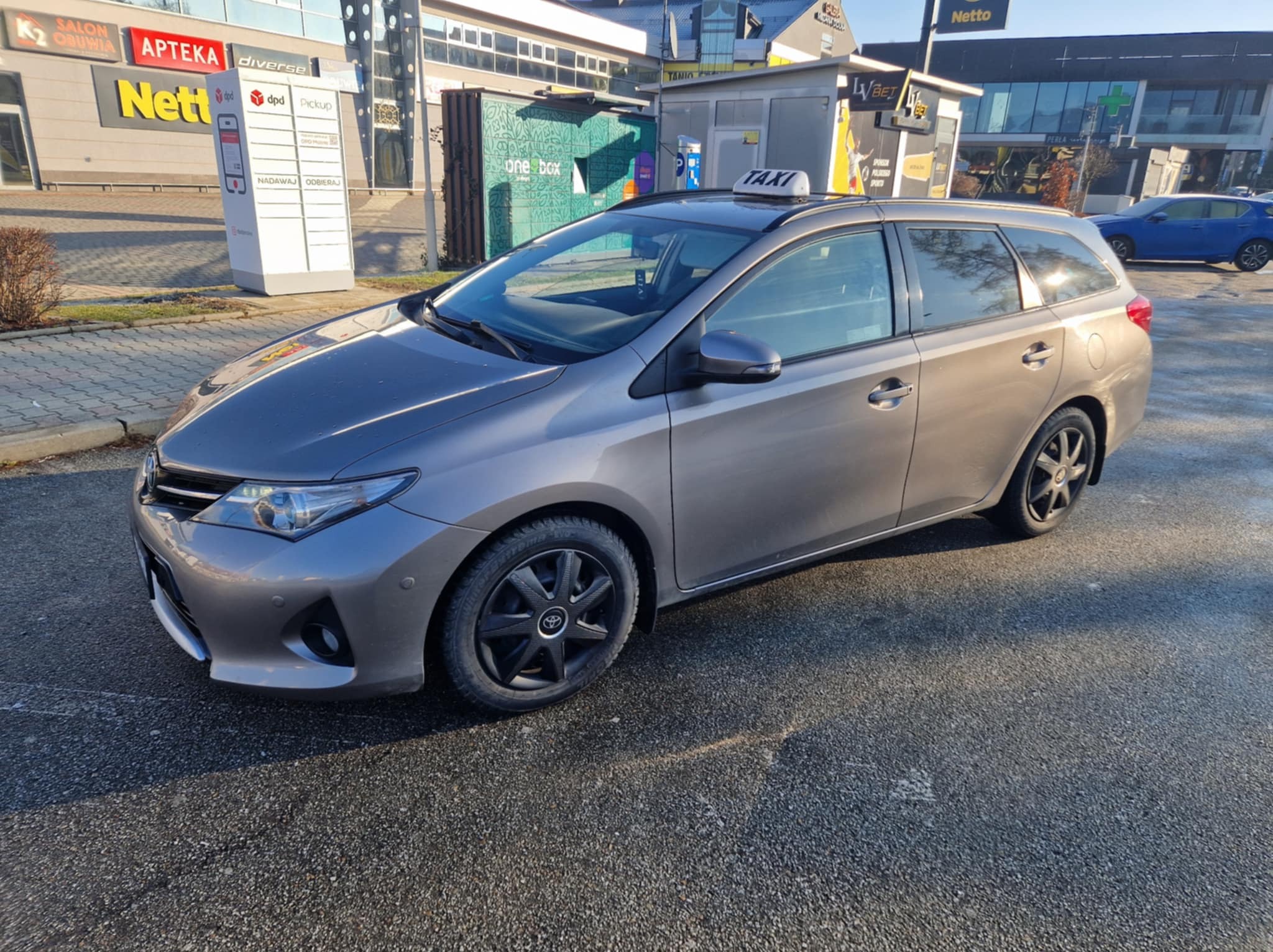 Szary samochód typu kombi z czarnymi felgami i napisem TAXI na dachu, zaparkowany na asfaltowym parkingu przed budynkami handlowymi z widocznymi szyldami sklepów i punktów usługowych.