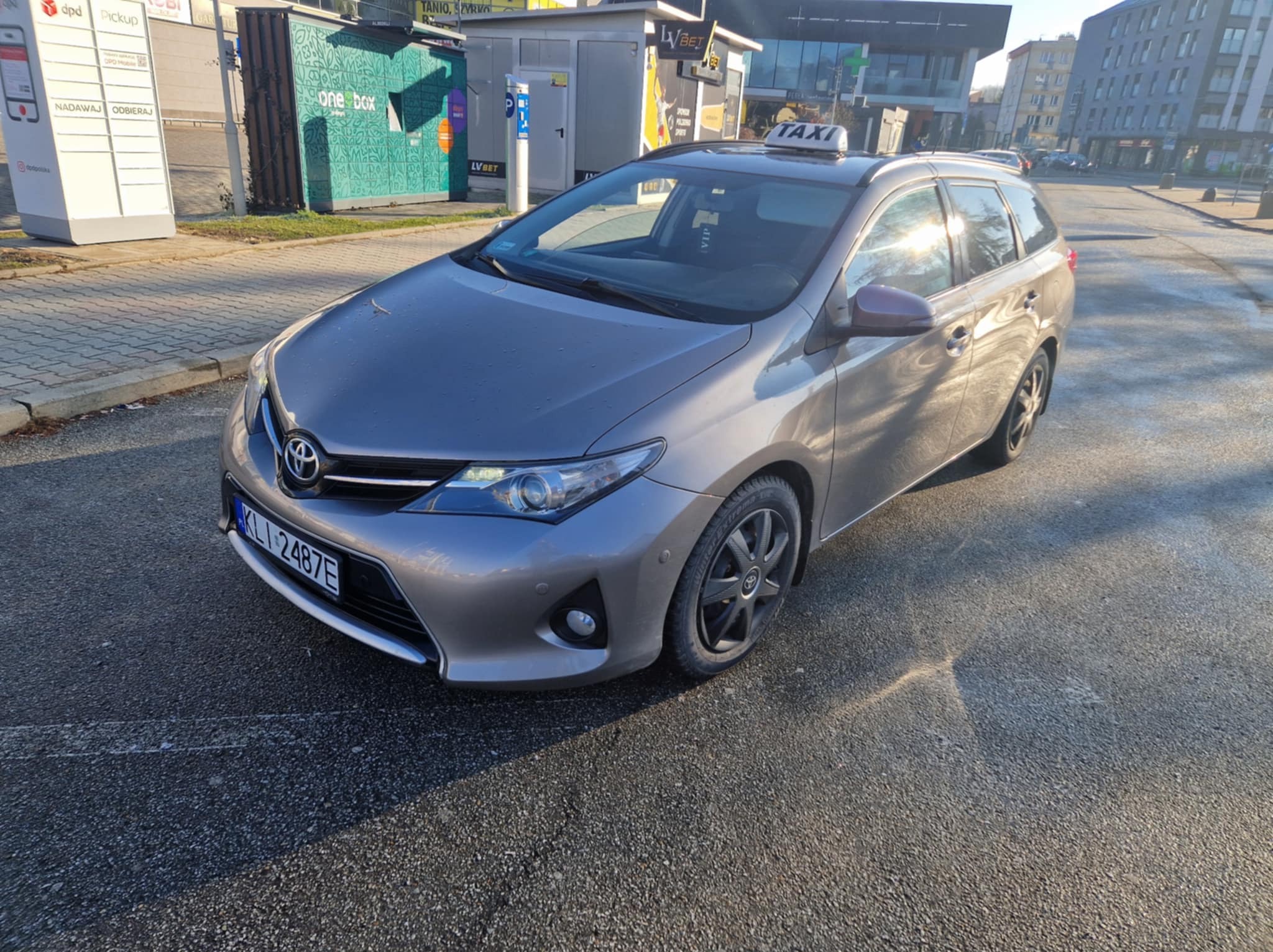 Szary samochód typu kombi z napisem TAXI na dachu, zaparkowany na ulicy w słoneczny dzień, widoczny automat paczkowy DPD i budynek z reklamą LVBet w tle.