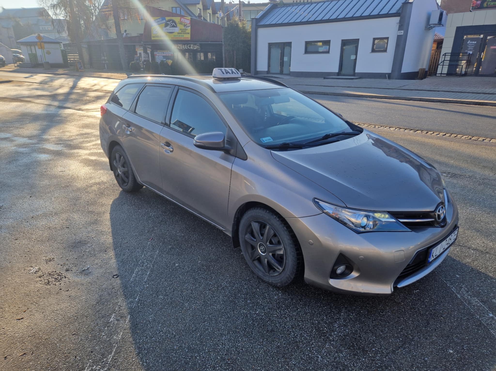 Szary samochód kombi z napisem TAXI na dachu, zaparkowany na ulicy w słoneczny dzień, z widocznymi budynkami w tle.