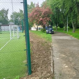 Smart Solutions Katarzyna Darewska - Sztuczna trawa boiska za siatką, obok ścieżka i zaparkowany samochód. Drzewa w tle. Dzień pochmurny.