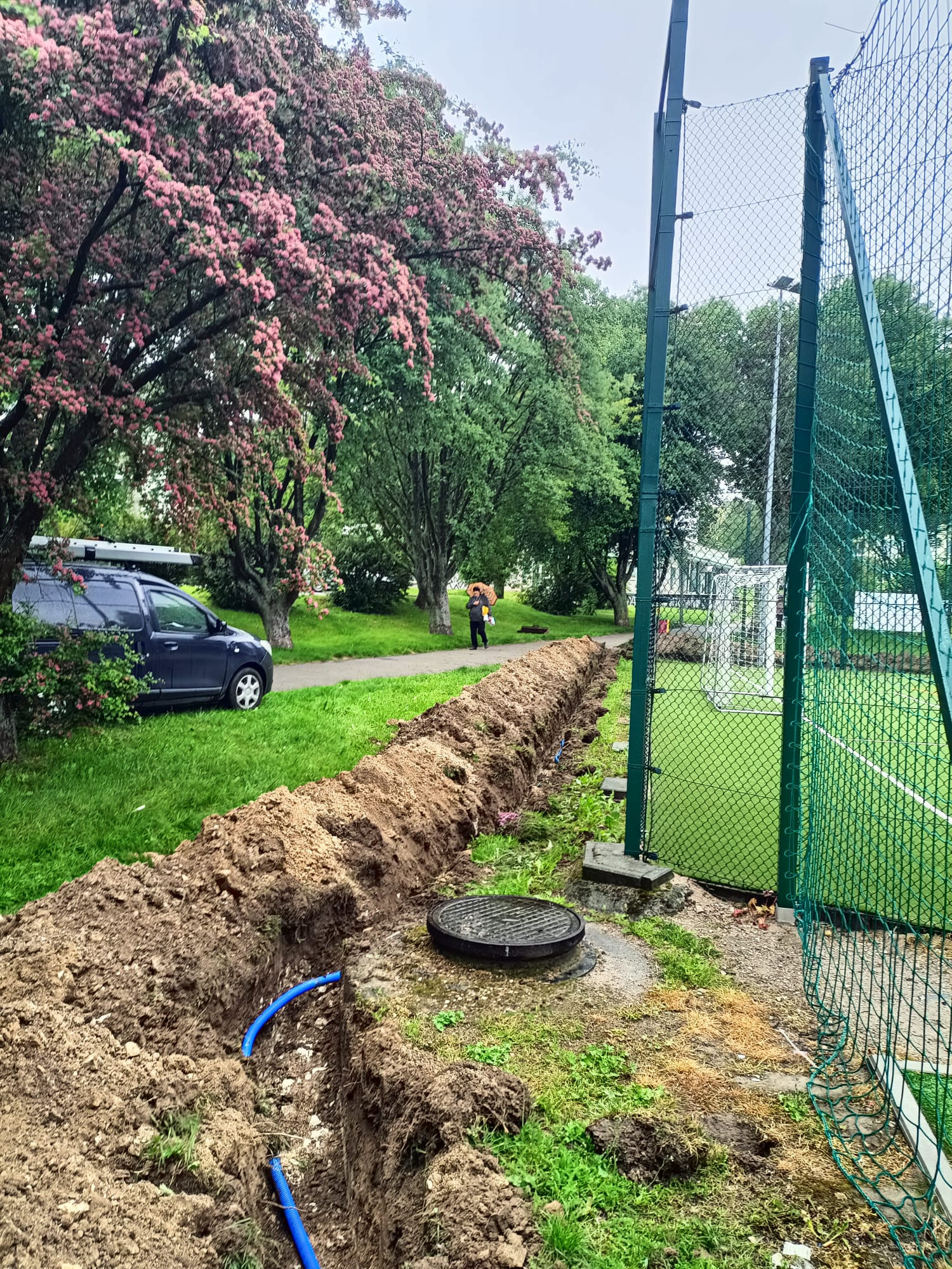 Wykop z niebieskimi rurami w ziemi, obok studzienka kanalizacyjna i siatka boiska sportowego. W tle kwitnące drzewo i zaparkowany samochód.