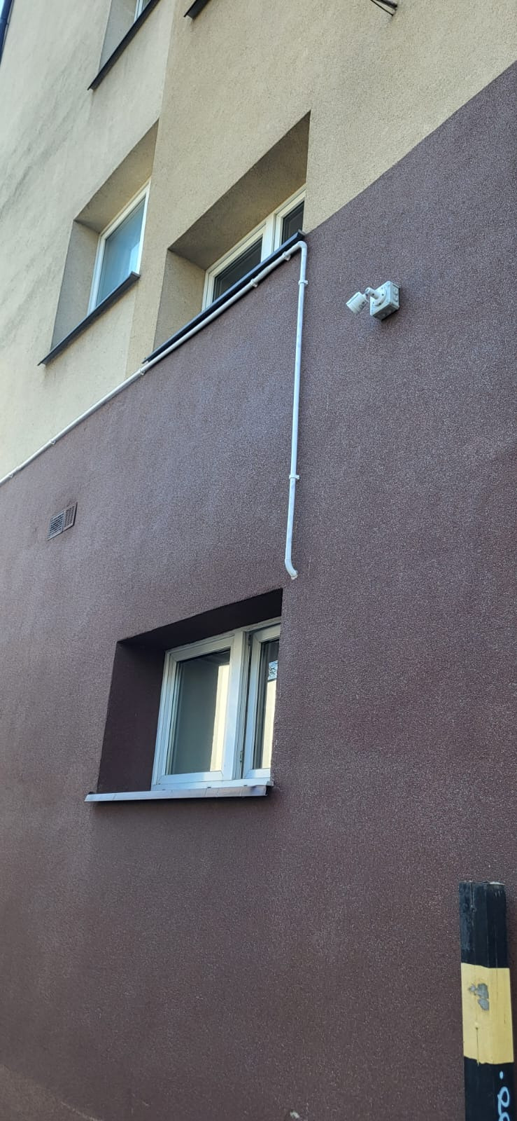 Brązowa elewacja budynku z oknami, białą rurą i zamontowaną lampą zewnętrzną. Ujęcie z dołu, widoczny fragment słupka z żółtymi elementami.