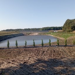 Firma Usługowo Handlowa Adrian Kuświk - Sztuczny zbiornik wodny otoczony ziemią, zabezpieczony folią i obsadzony młodymi tujami. W tle widoczny mur oporowy, drzewa i kukurydza.