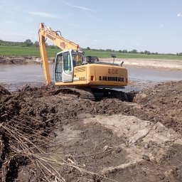 Firma Usługowo Handlowa Adrian Kuświk - Żółta koparka Liebherr 900 na podmokłym terenie z błotem i gałęziami, w tle zielone pola i błękitne niebo.