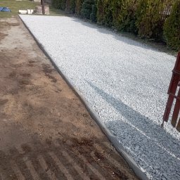 Firma Usługowo Handlowa Adrian Kuświk - Utwardzona droga z jasnego kruszywa, obrzeża z betonowych krawężników, widok na ogród z tujami i studnią z figurką bociana.