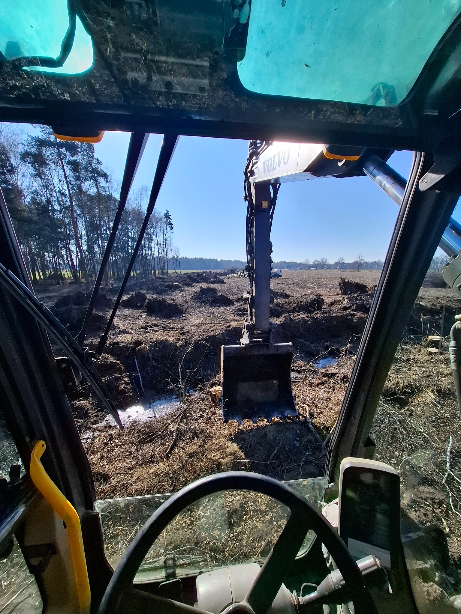 Widok z kabiny koparki Volvo na teren po wycince drzew, z widocznymi korzeniami i świeżo naruszoną ziemią, w tle las i błękitne niebo.