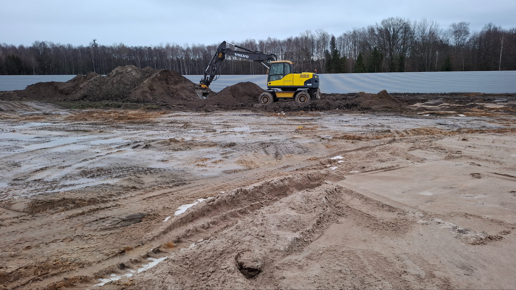 Żółta koparka Volvo na błotnistym placu budowy, z kupami ziemi i metalowym ogrodzeniem w tle, pochmurne niebo.