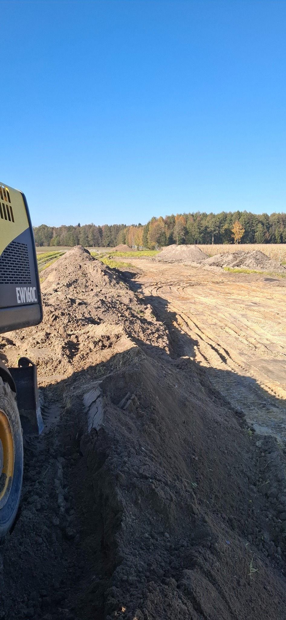 Żółta koparka EW160C częściowo widoczna, obok pryzma ziemi na wyrównanym terenie z widocznymi śladami opon i drzewami w tle, błękitne niebo.