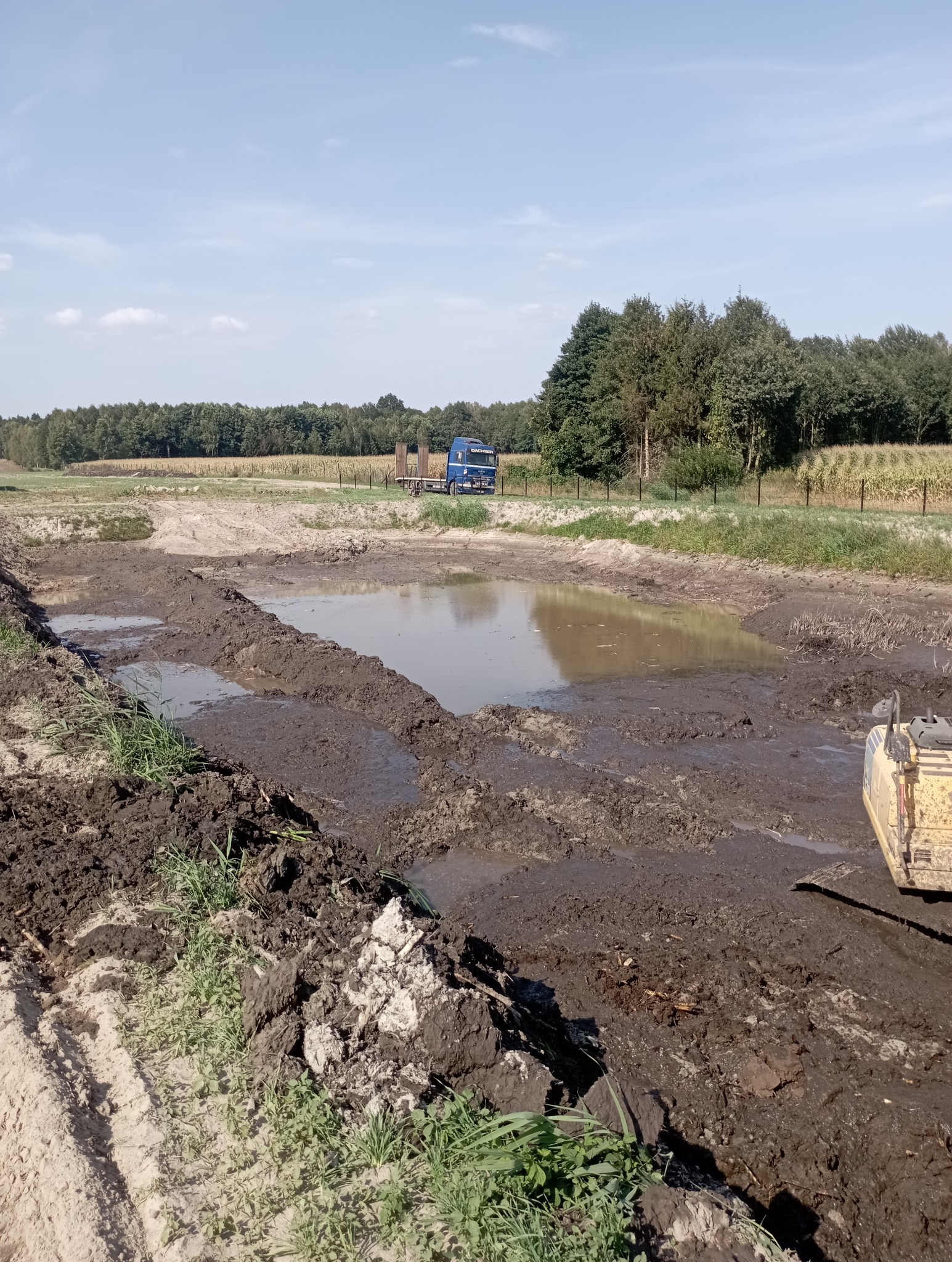 Wykop pod zbiornik wodny wypełniony wodą, na drugim planie niebieska ciężarówka na tle drzew