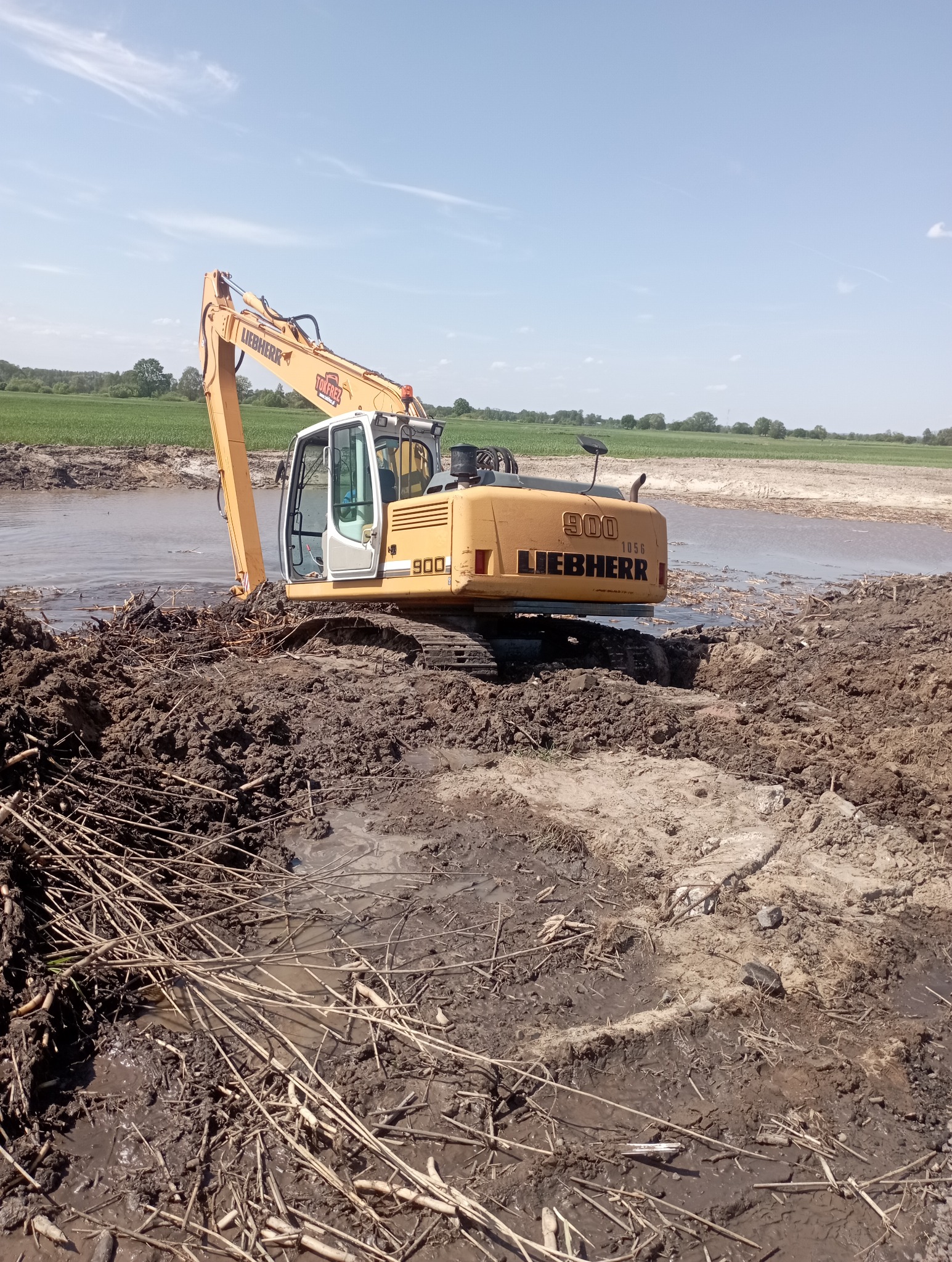 Żółta koparka Liebherr 900 na podmokłym terenie z błotem i gałęziami, w tle zielone pola i błękitne niebo.