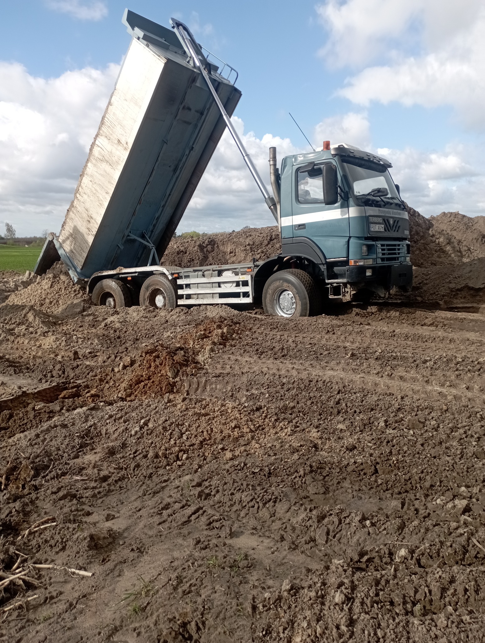 Ciężarówka wywrotka z podniesioną skrzynią ładunkową, częściowo zanurzona w grząskim, błotnistym terenie. Widoczne ślady opon i świeżo usypana ziemia.