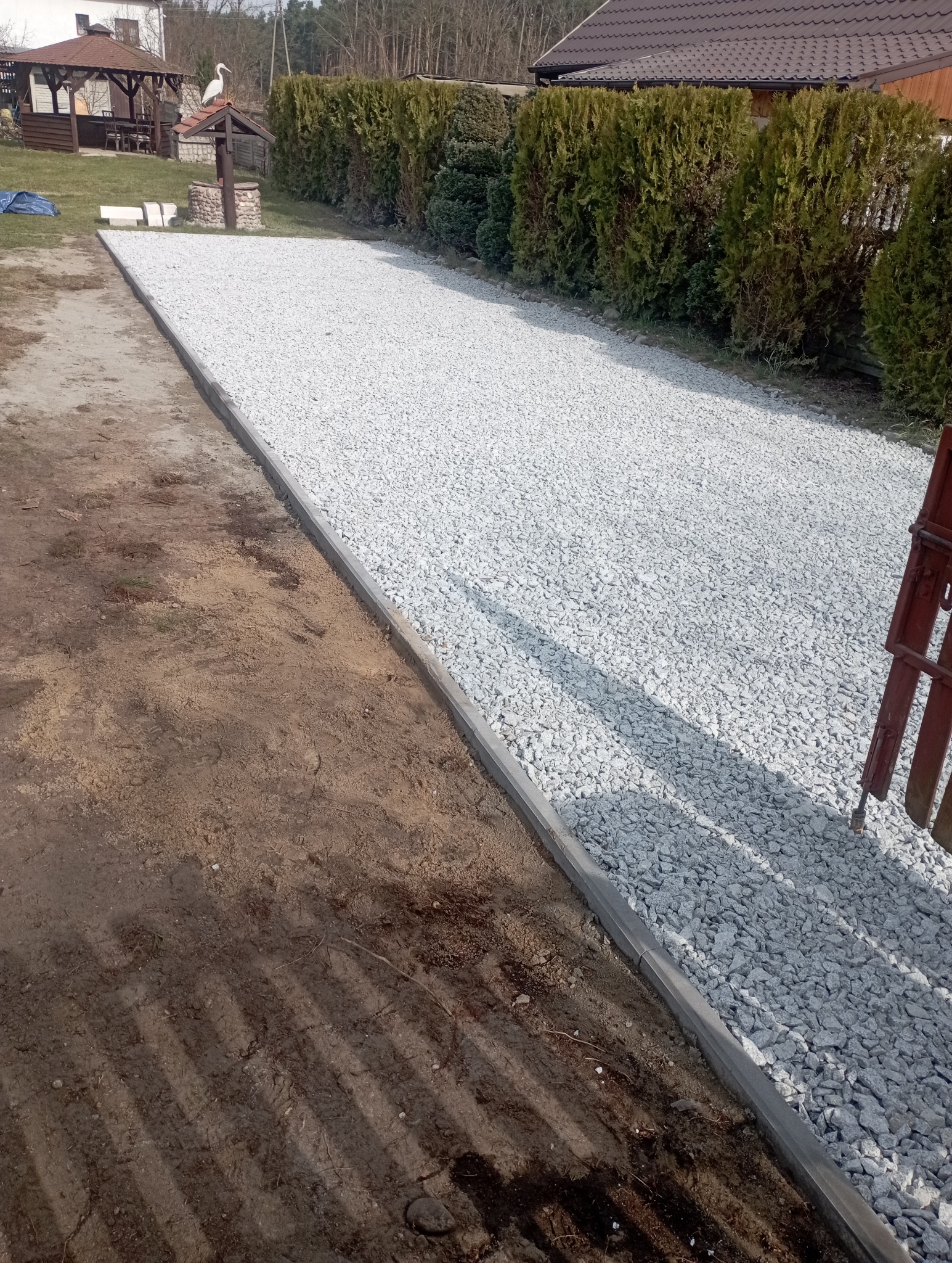 Utwardzona droga z jasnego kruszywa, obrzeża z betonowych krawężników, widok na ogród z tujami i studnią z figurką bociana.