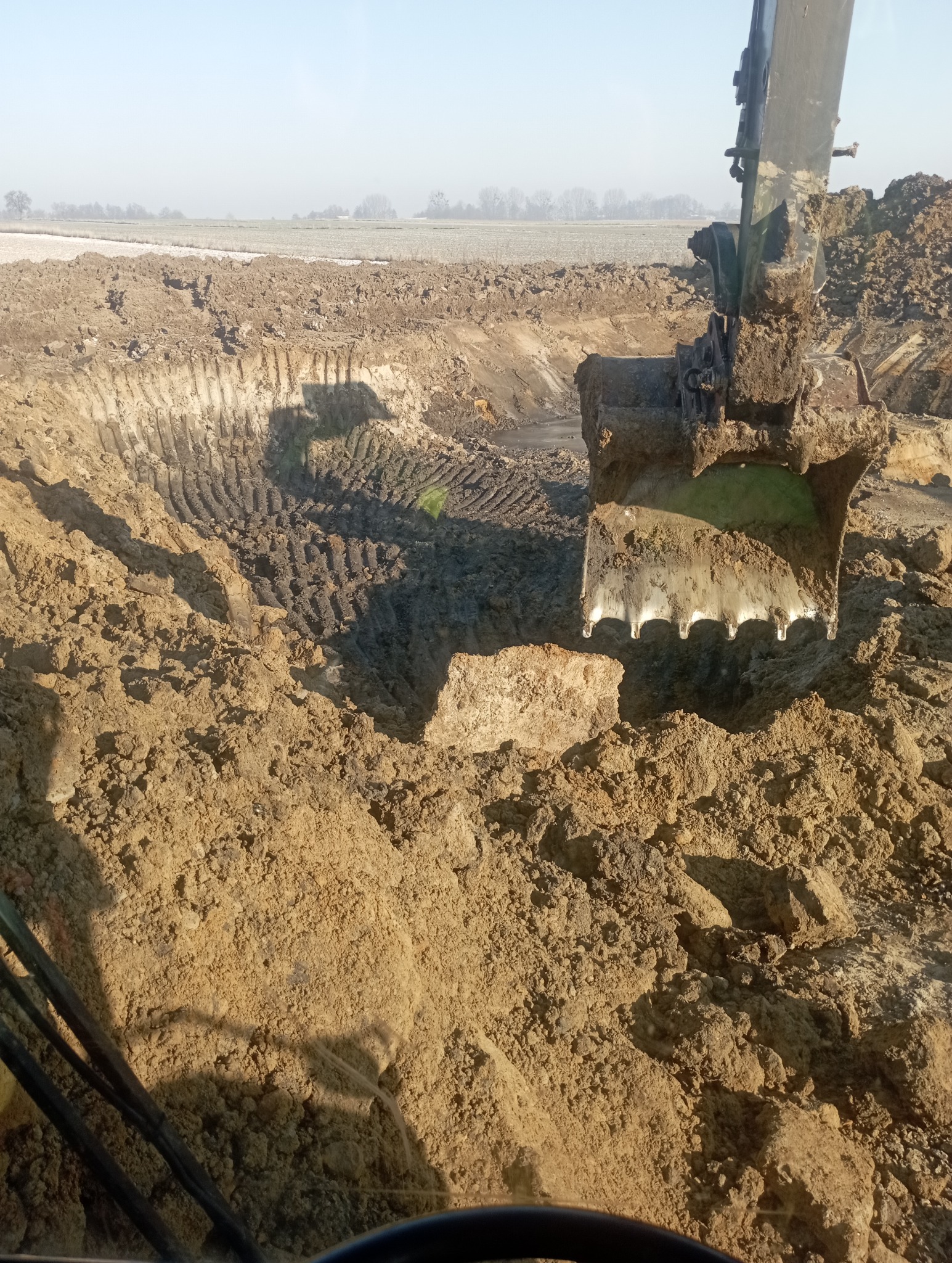 Ziemia wykopana przez łyżkę koparki, widok z kabiny operatora, w tle krajobraz pola w lekkiej mgle.