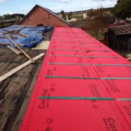 Częściowo zdjęty dach starego domu, pokryty czerwoną membraną dachową Corotop Red Strong, zabezpieczoną zielonymi listwami, z widoczną konstrukcją więźby i fragmentem niebieskiej plandeki