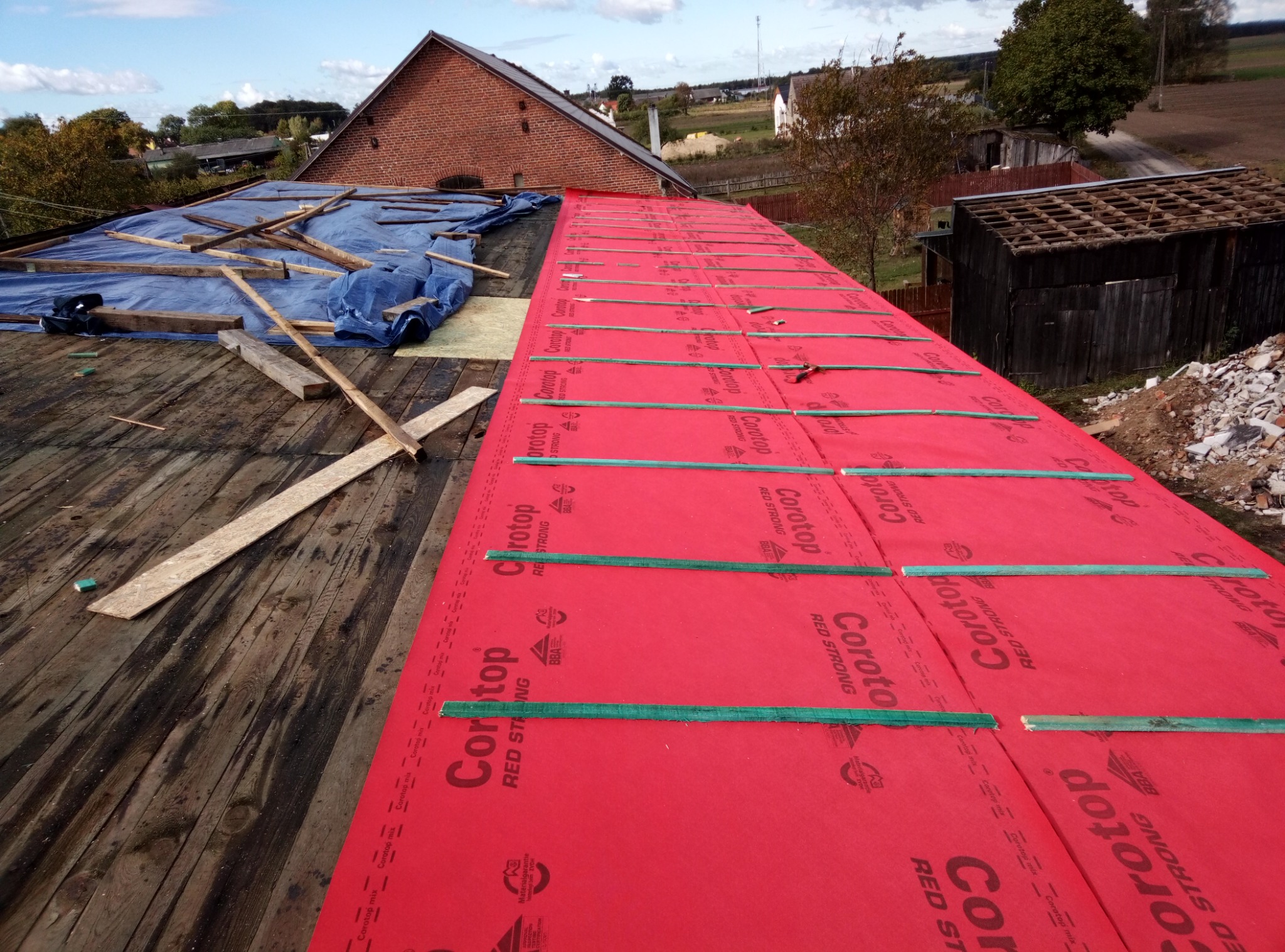 Częściowo zdjęty dach starego domu, pokryty czerwoną membraną dachową Corotop Red Strong, zabezpieczoną zielonymi listwami, z widoczną konstrukcją więźby i fragmentem niebieskiej plandeki