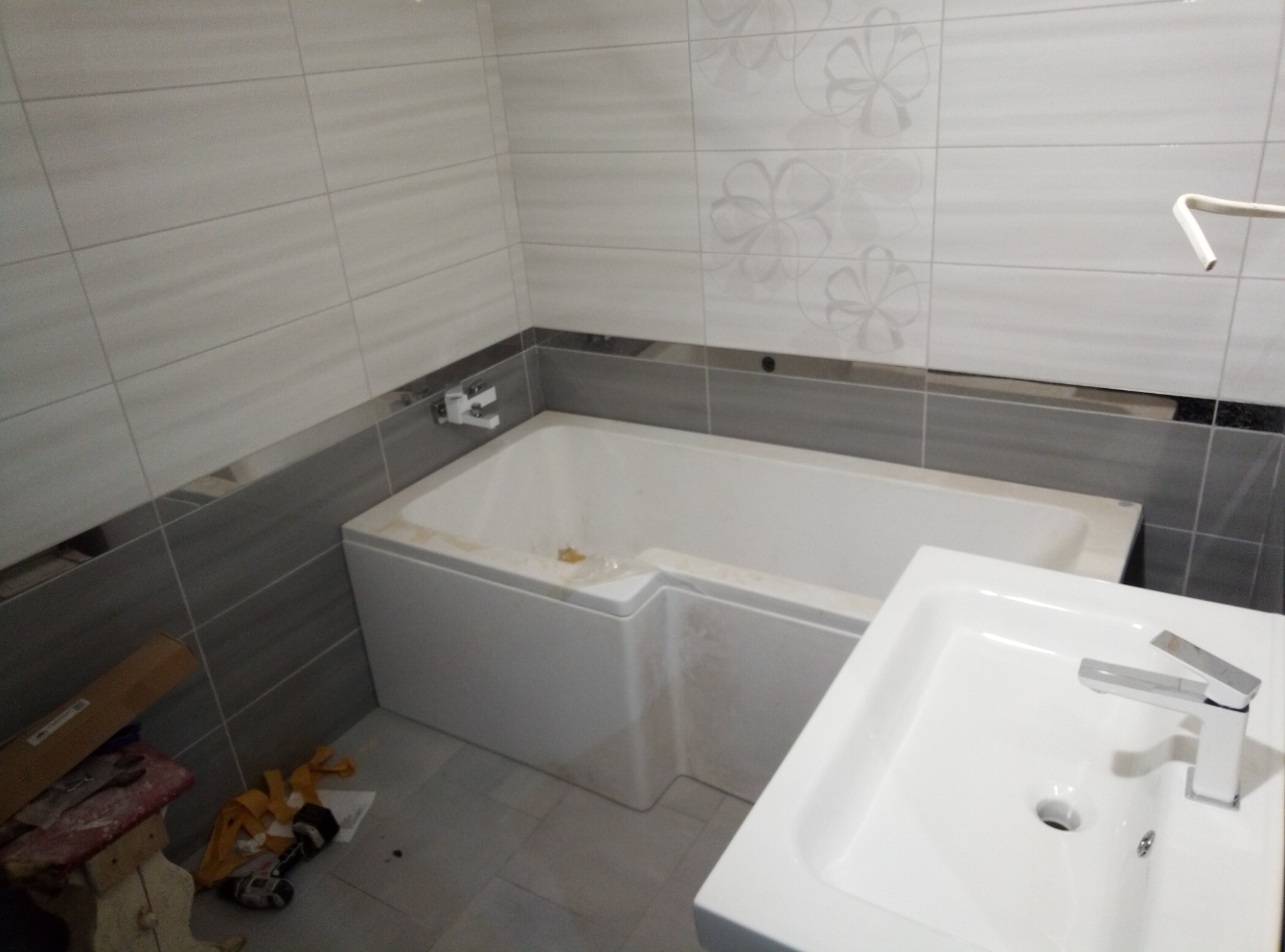 Niedokończona łazienka z wanną, umywalką i szarymi płytkami z kwiatowym wzorem, widoczne narzędzia i materiały budowlane w rogu.