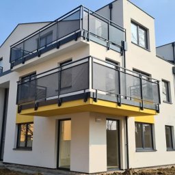 Balu-Inwest - Nowoczesny budynek wielorodzinny z balkonami zabezpieczonymi czarnymi, siatkowymi barierkami, żółte akcenty kolorystyczne na elewacji, widoczne świeżo posadzone tuje.