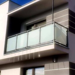 Balu-Inwest - Nowoczesny balkon z szklaną balustradą i metalową ramą, widok z dołu na tle elewacji budynku z elementami dekoracyjnymi.