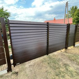 Balu-Inwest - Nowoczesne, brązowe ogrodzenie panelowe z poziomych listew, częściowo otwarte, z widocznym mechanizmem automatyki bramy, na tle trawnika i zabudowań jednorodzinnych.