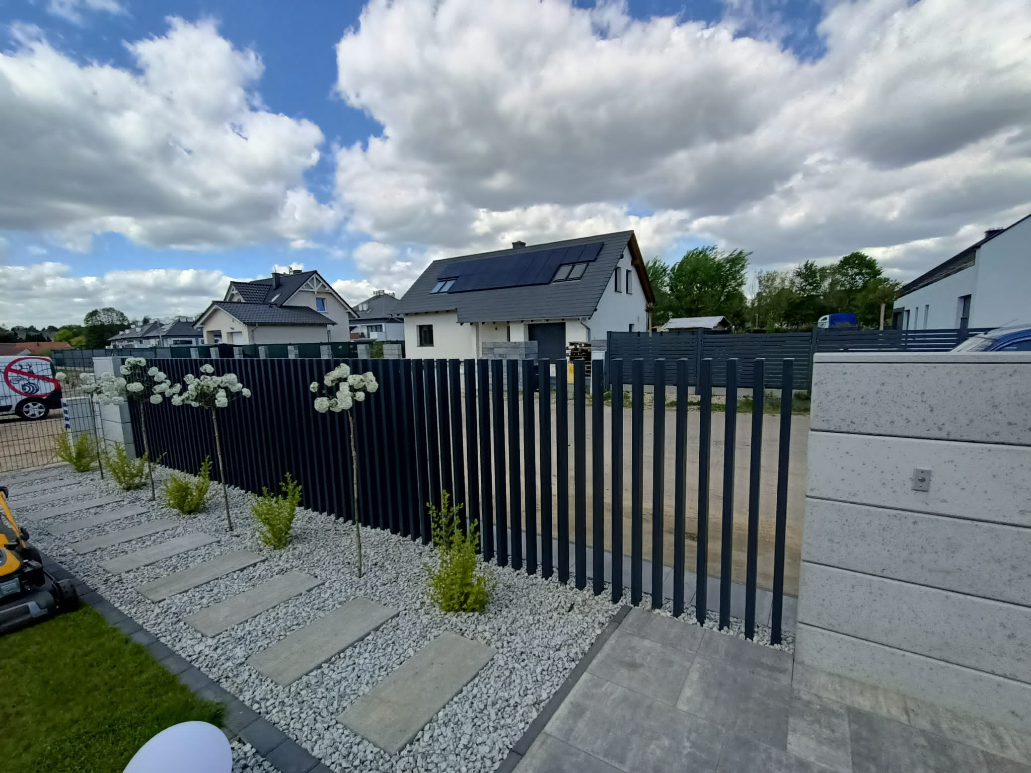 Nowoczesne ogrodzenie panelowe w kolorze antracytowym z pionowymi sztachetami, oddzielające posesję z domem z panelami słonecznymi, ozdobione kulistymi drzewkami i jasnymi kamieniami.