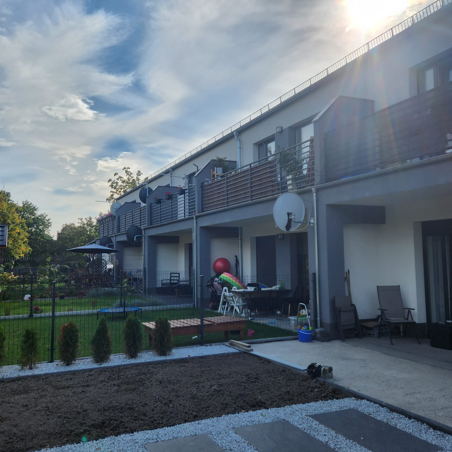 Rząd balkonów z poziomymi, metalowymi barierkami w szeregowej zabudowie, widok na ogród z młodymi tujami i placem zabaw.