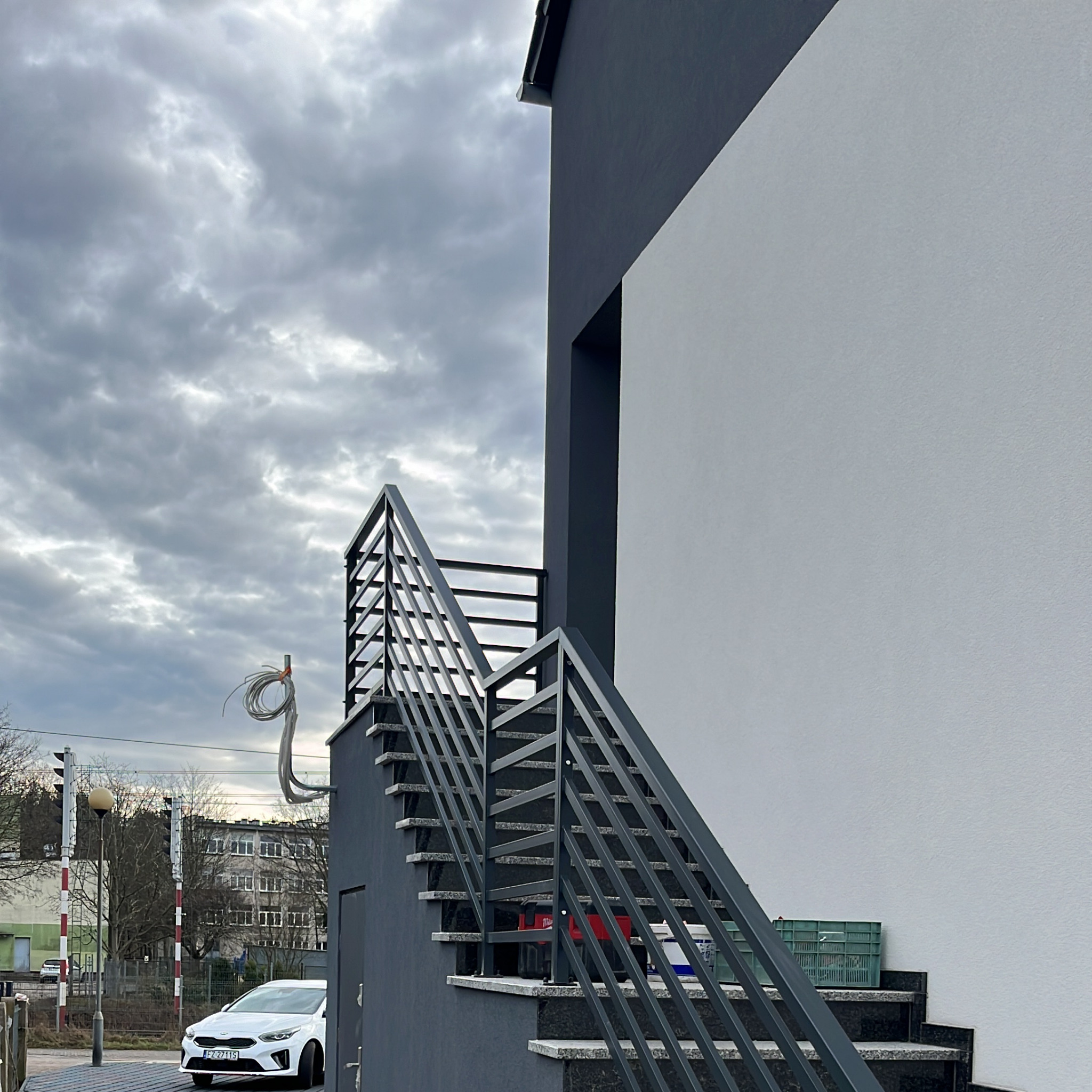 Zewnętrzne schody z grafitową metalową balustradą o prostym, nowoczesnym designie, prowadzące do budynku o białej elewacji, widoczne narzędzia i pojemniki na schodach, pochmurne niebo w tle.