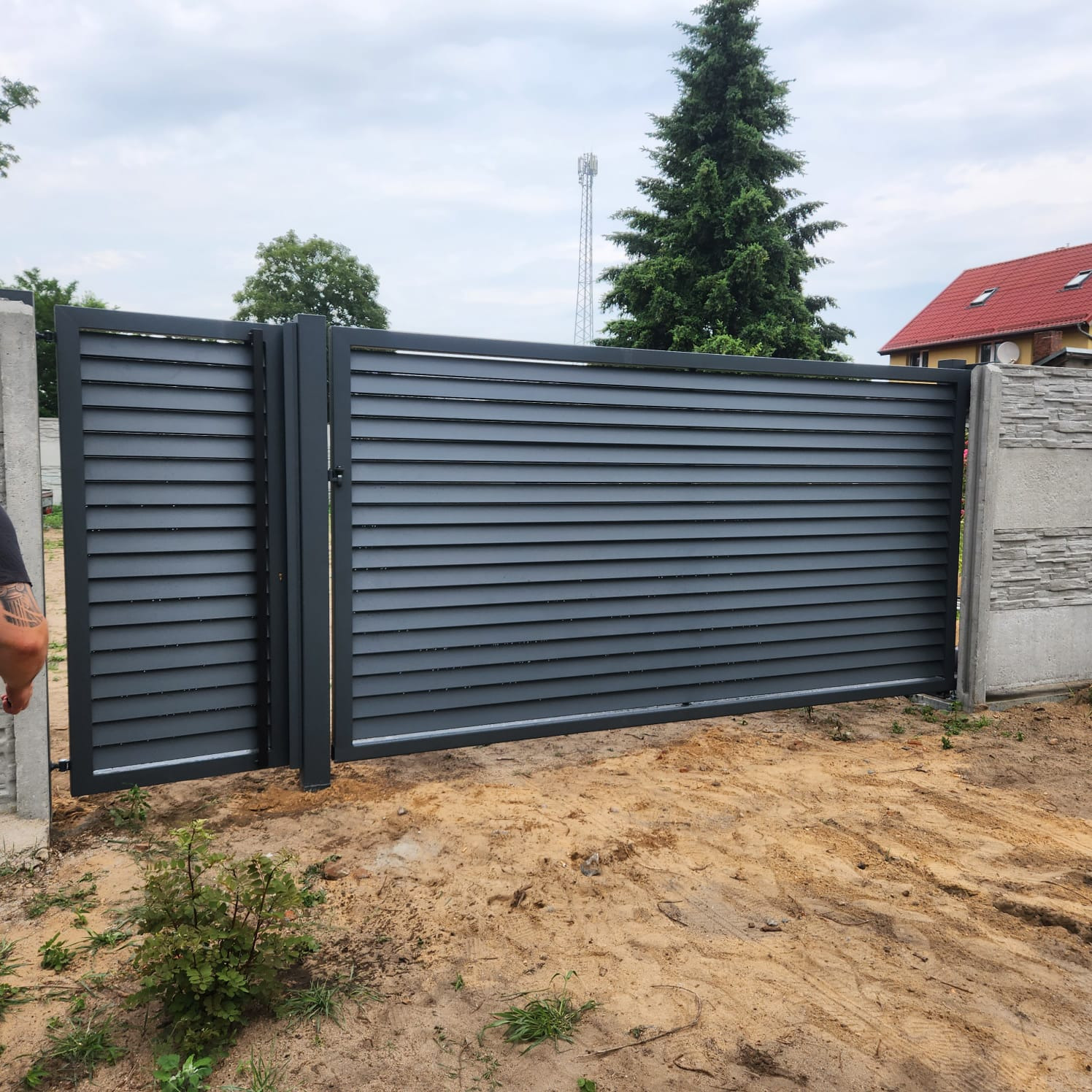 Nowoczesna, pozioma brama lamelowa w kolorze grafitowym, zamontowana między betonowymi słupkami z dekoracyjnym wzorem, na tle piaszczystego gruntu i zieleni. Widoczna część ręki osoby...
