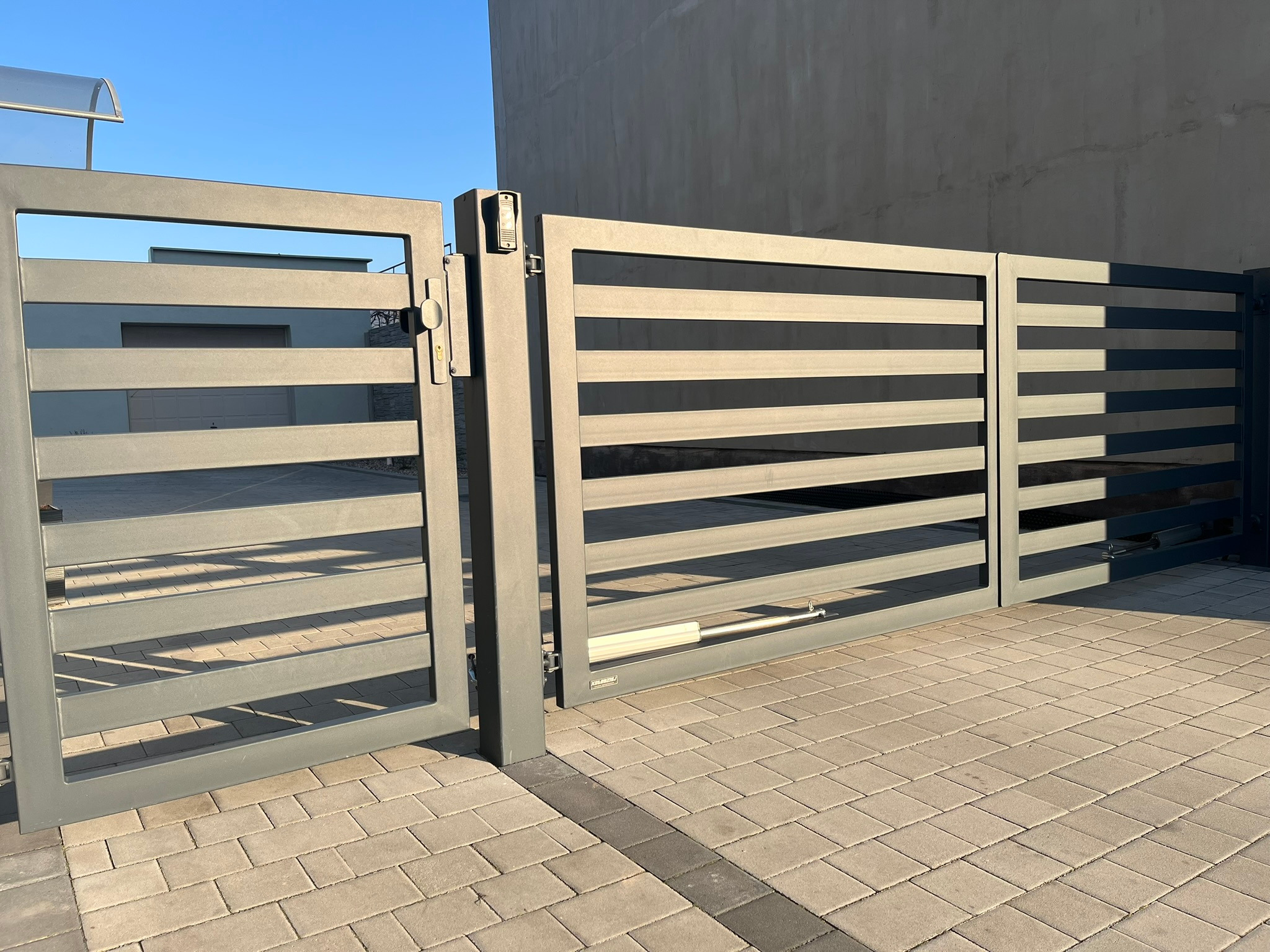 Nowoczesna, szara brama przesuwna z poziomymi panelami, częściowo otwarta, z widocznym mechanizmem automatycznego otwierania, na tle kostki brukowej i szarego budynku.