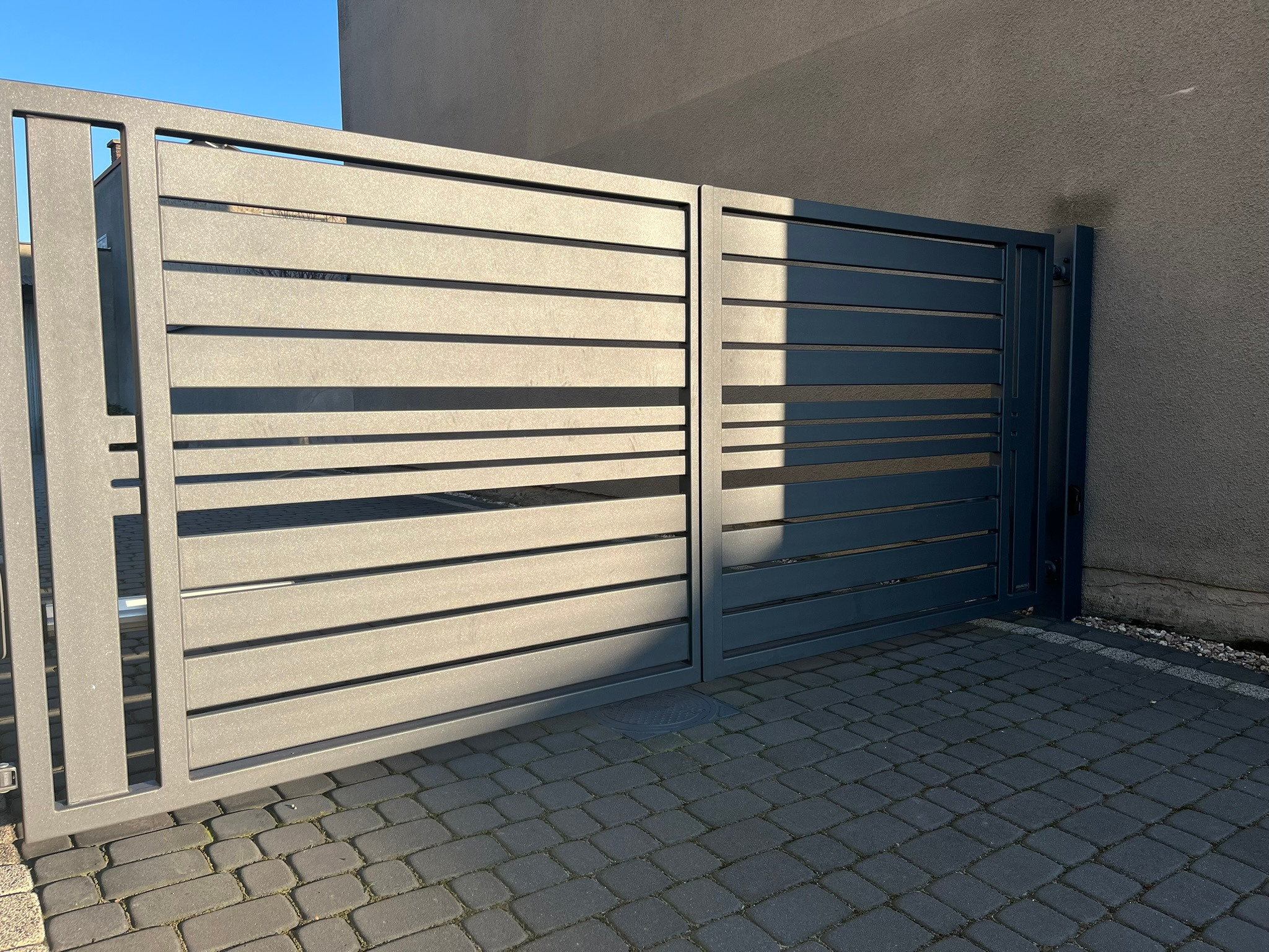 Nowoczesna, metalowa brama przesuwna z poziomymi panelami, częściowo oświetlona słońcem, na tle szarej ściany i kostki brukowej.