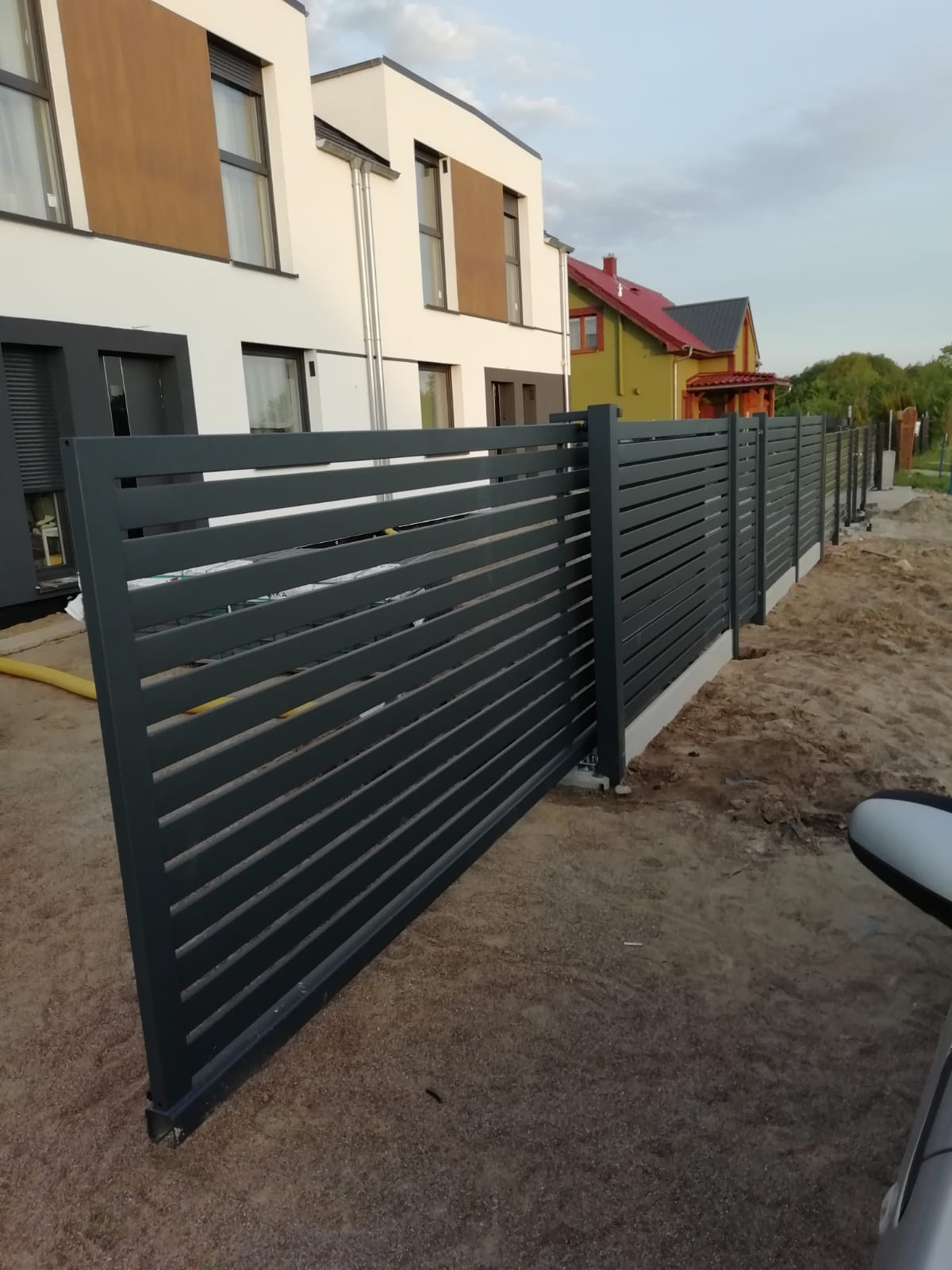 Nowoczesne ogrodzenie posesji z poziomych, szarych profili, widok z boku na tle domu z elementami brązowymi i zielonego domu z czerwonym dachem.