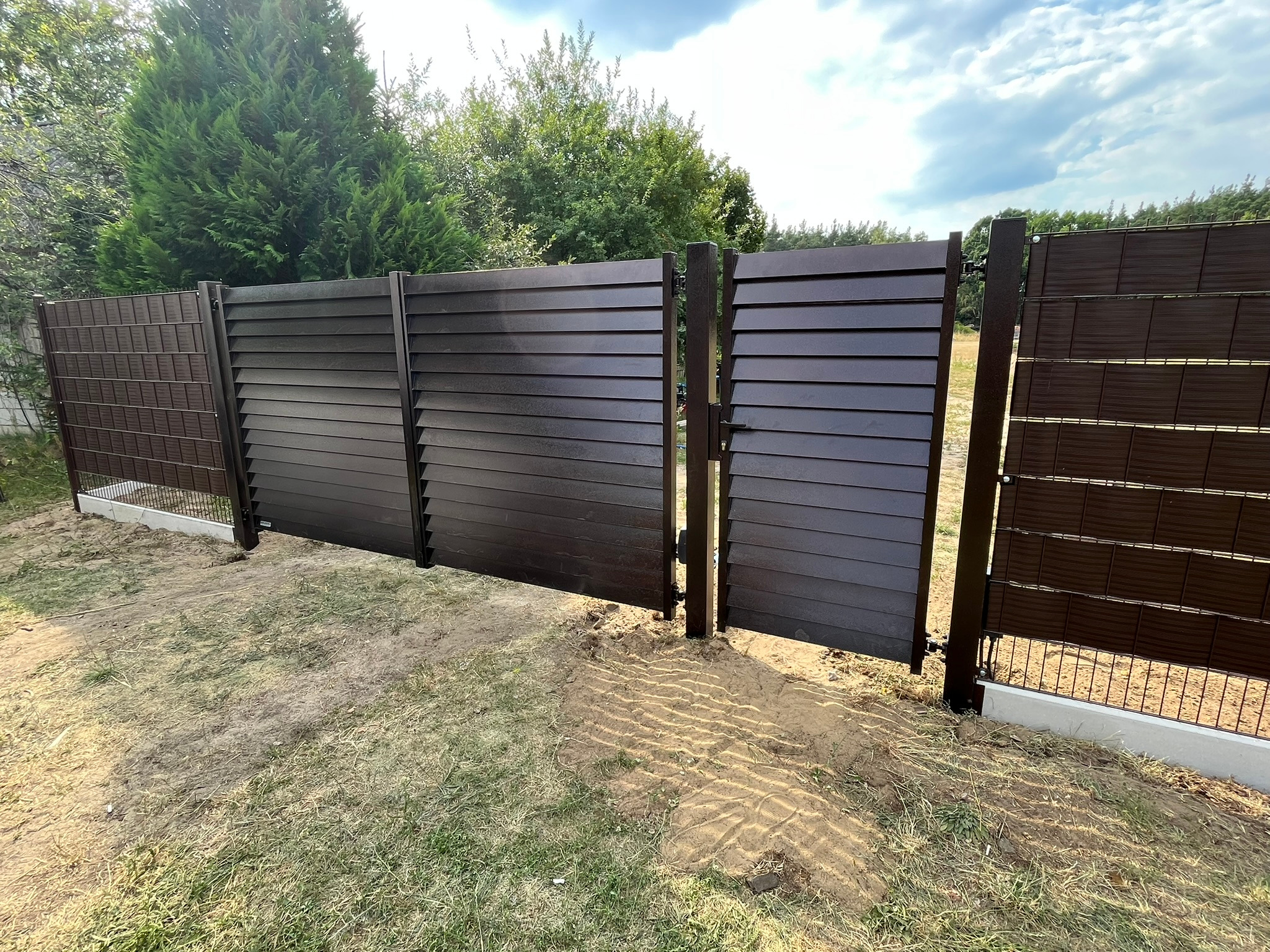 Brązowe, poziome panele ogrodzeniowe z furtką, częściowo ażurowe, na tle zieleni drzew i błękitnego nieba. Widoczne świeże ślady prac ziemnych.