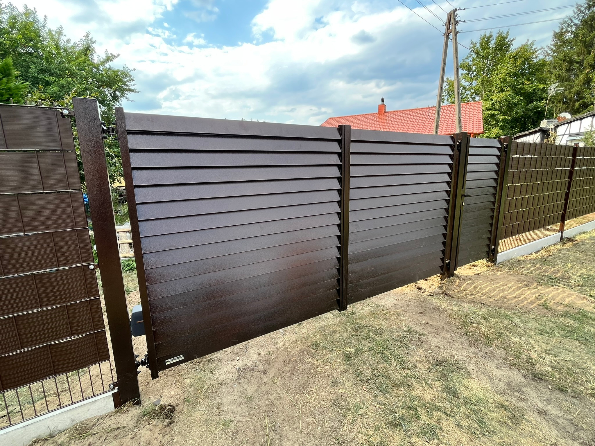 Nowoczesne, brązowe ogrodzenie panelowe z poziomych listew, częściowo otwarte, z widocznym mechanizmem automatyki bramy, na tle trawnika i zabudowań jednorodzinnych.