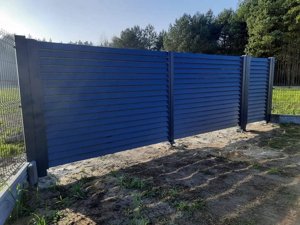 Nowoczesne, poziome ogrodzenie lamelowe w kolorze antracytowym, zlokalizowane w Zbąszyniu, z widoczną siatką ogrodzeniową w tle i niezagospodarowanym terenem u podstawy.