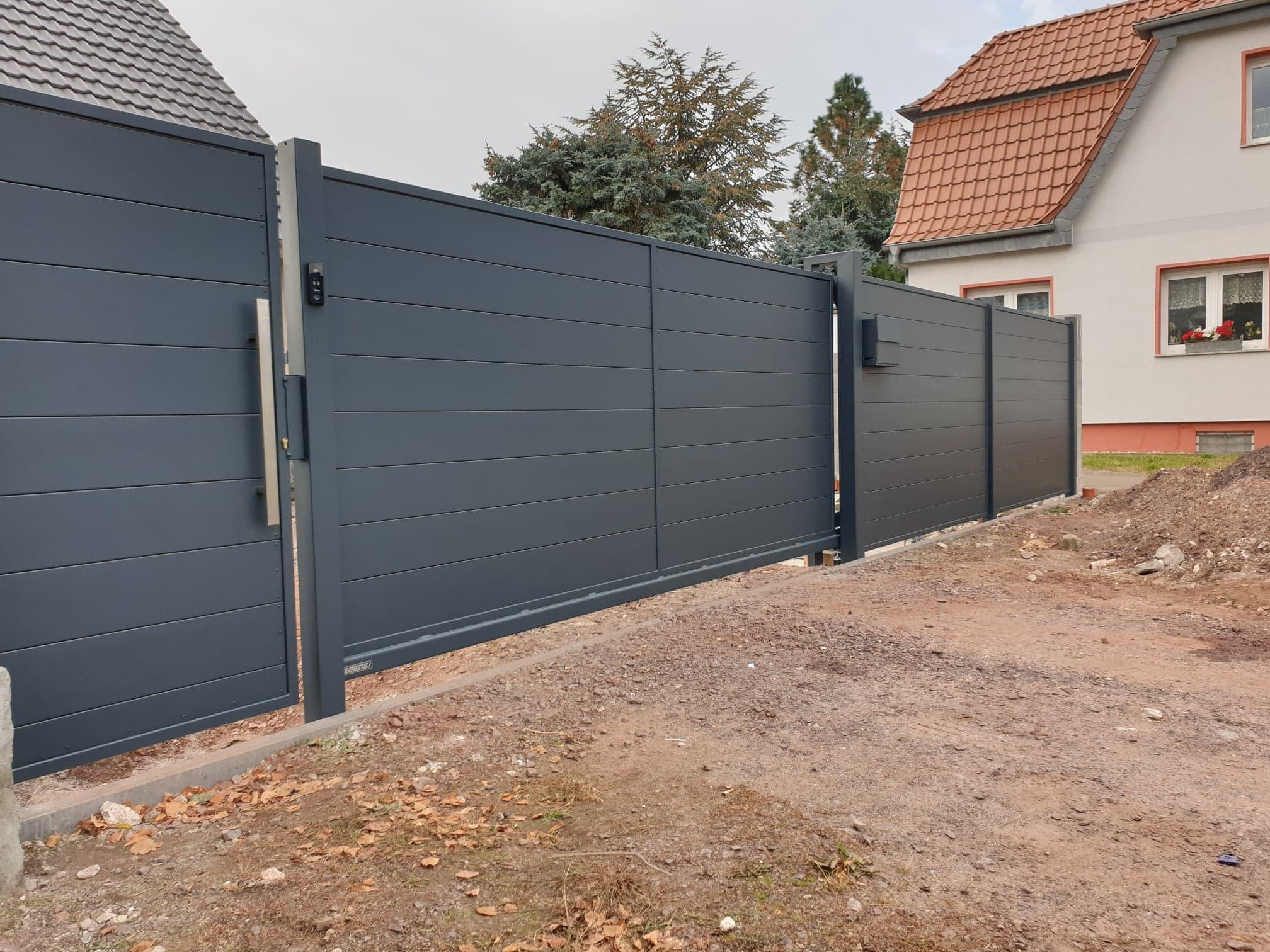 Nowoczesna, przesuwna brama w kolorze grafitowym z poziomymi panelami, wyposażona w zamek elektroniczny, na tle domu z czerwonym dachem i drzew iglastych.