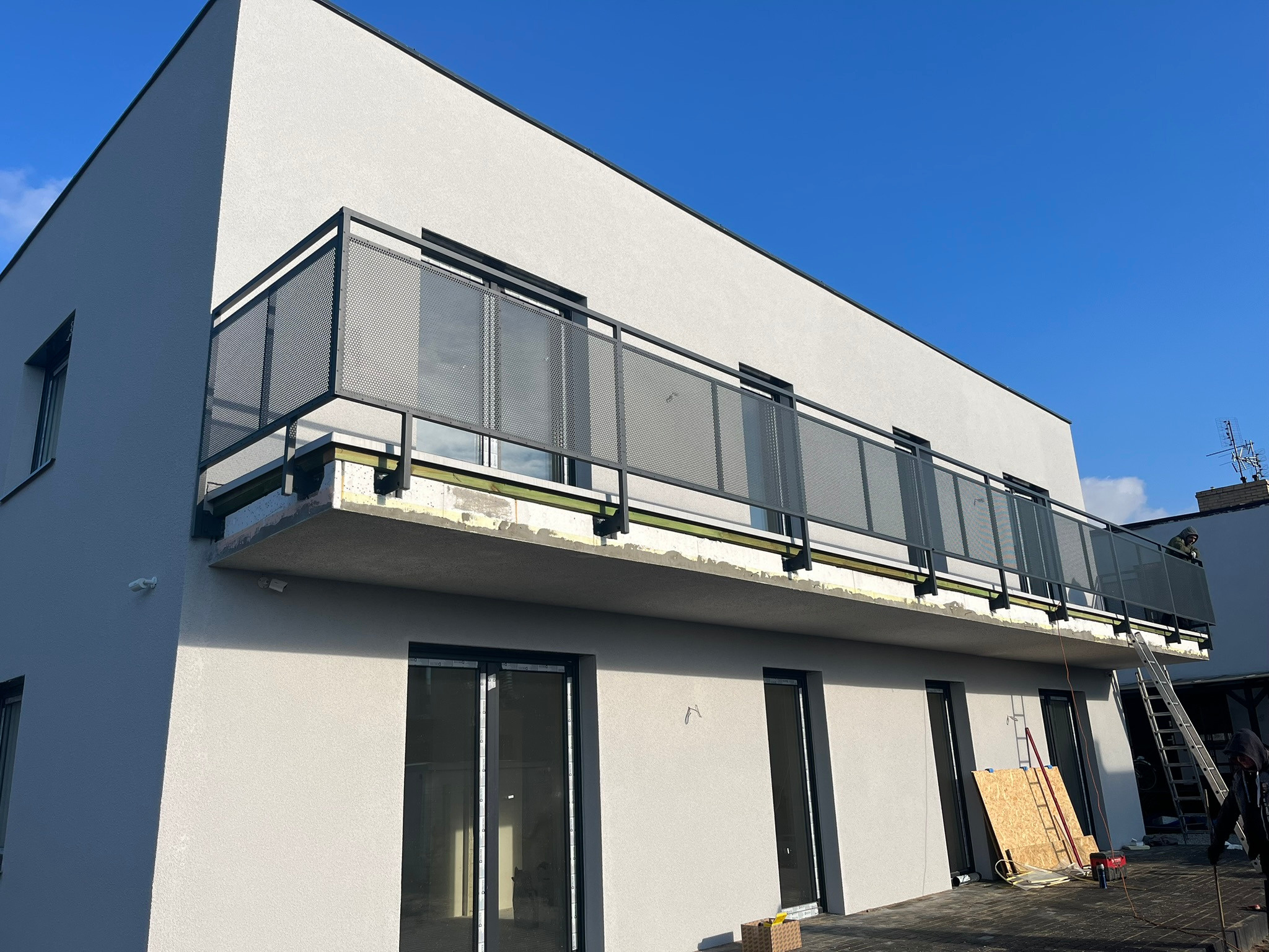 Nowoczesny balkon z metalową ażurową balustradą w trakcie montażu, widoczne narzędzia i materiały budowlane, jasna elewacja budynku.