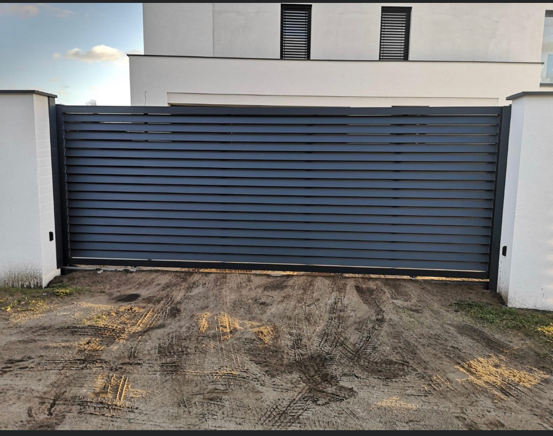 Nowoczesna, przesuwna brama posesyjna z poziomych, szarych lameli, osadzona między białymi słupkami, z widocznymi śladami opon na ziemi przed bramą.