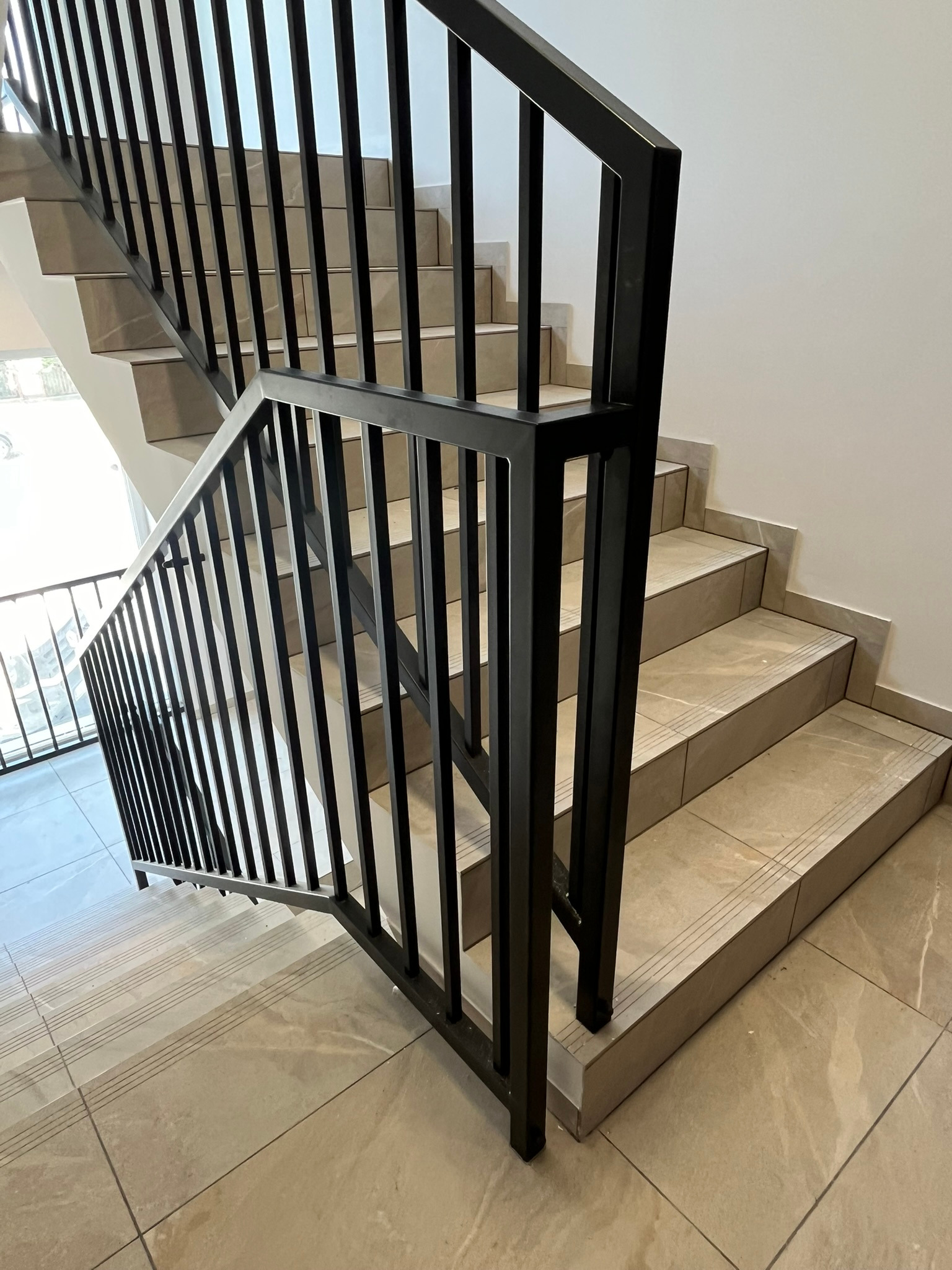 Nowoczesna, czarna balustrada z pionowych prętów, zamontowana wzdłuż schodów wyłożonych beżowymi płytkami imitującymi marmur, widok z góry.