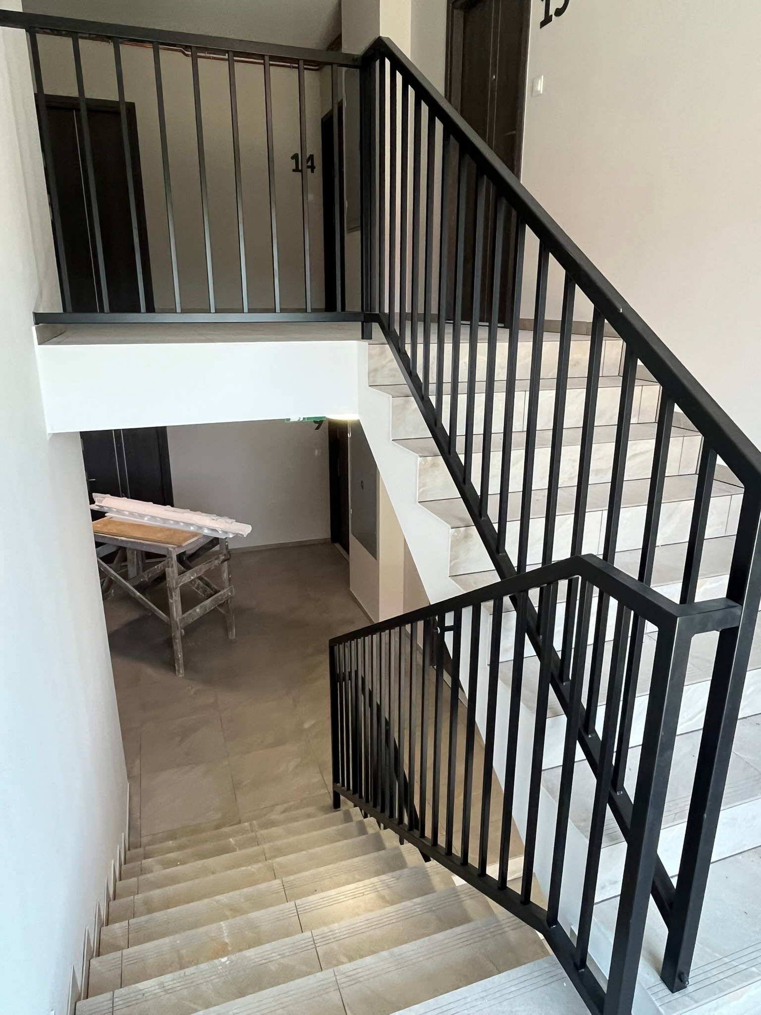 Schody wykończone jasnymi płytkami z czarną, metalową balustradą pionową; w tle widoczny stół roboczy i drzwi do mieszkań.