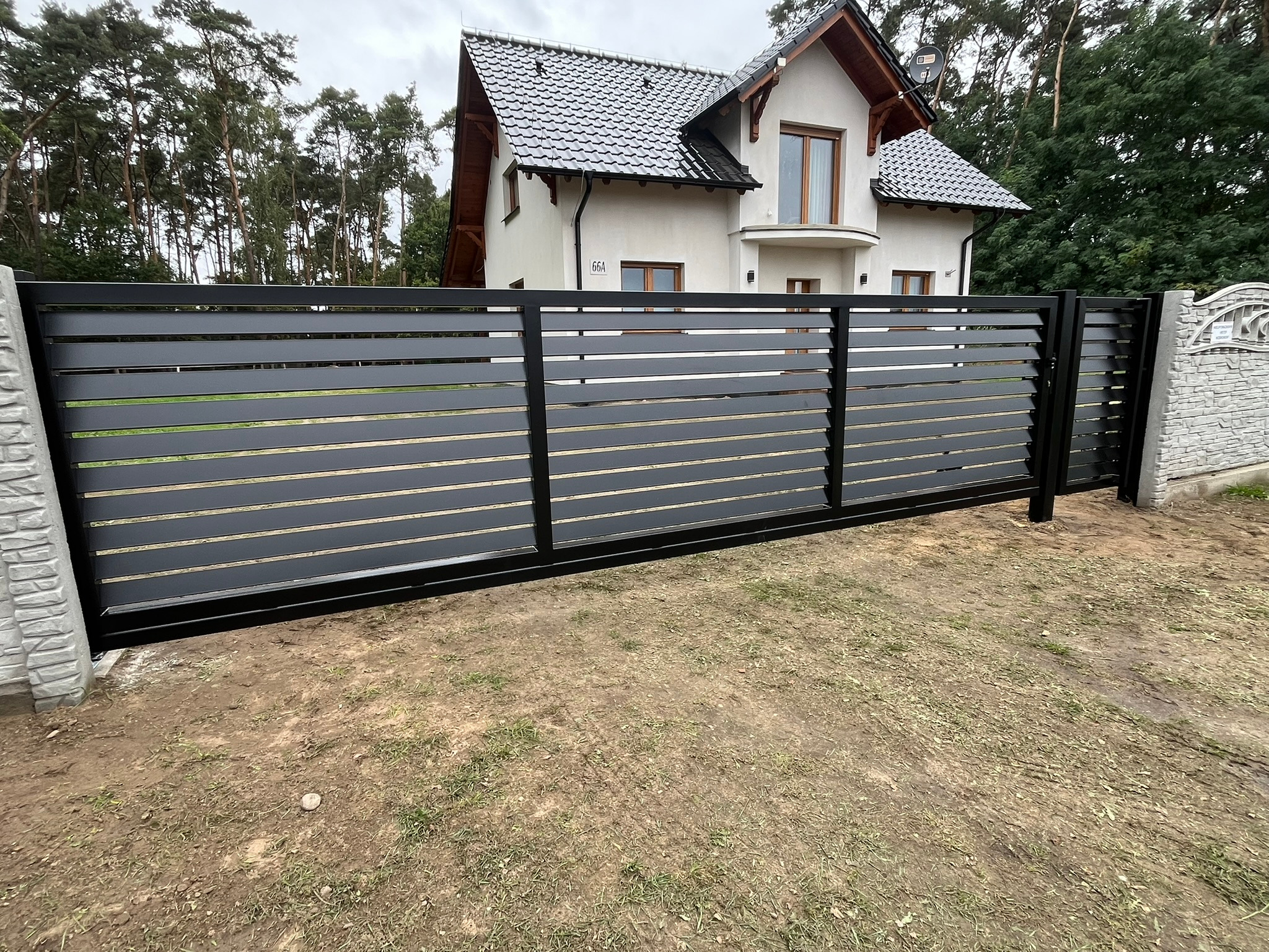 Nowoczesna, przesuwna brama w kolorze antracytowym z poziomymi panelami na tle domu z jasną elewacją i ciemną dachówką, otoczona zielenią.
