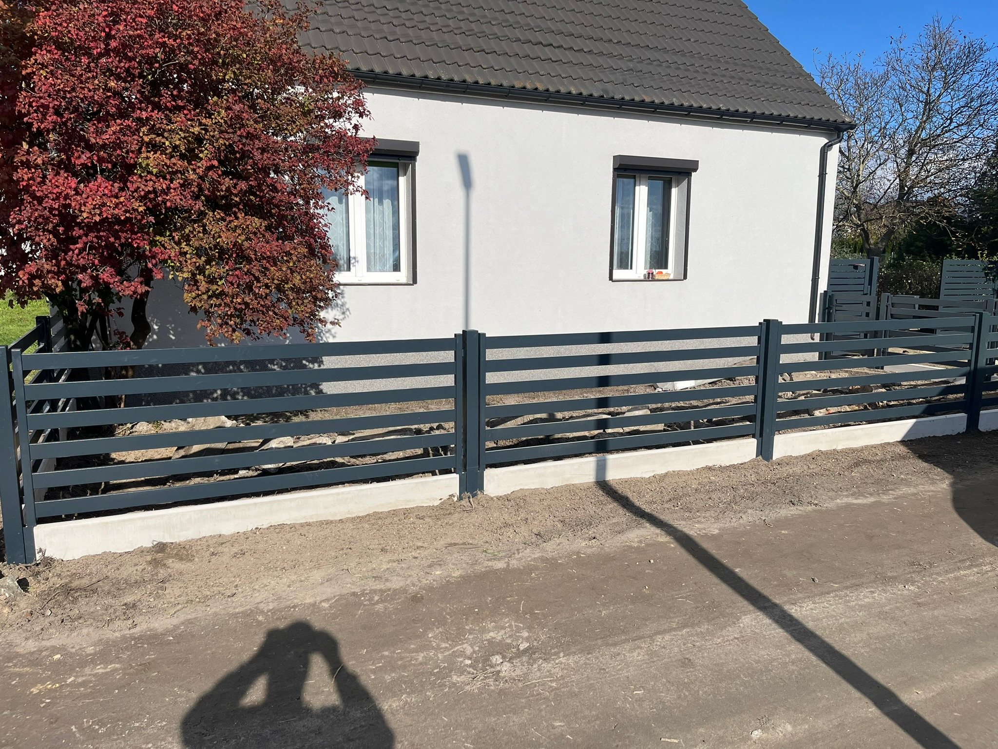 Nowoczesna, pozioma, metalowa balustrada w kolorze antracytowym, zamontowana na betonowej podmurówce przed domem z białymi ścianami i brązową dachówką, obok drzewo o czerwonych liściach, cień osoby...