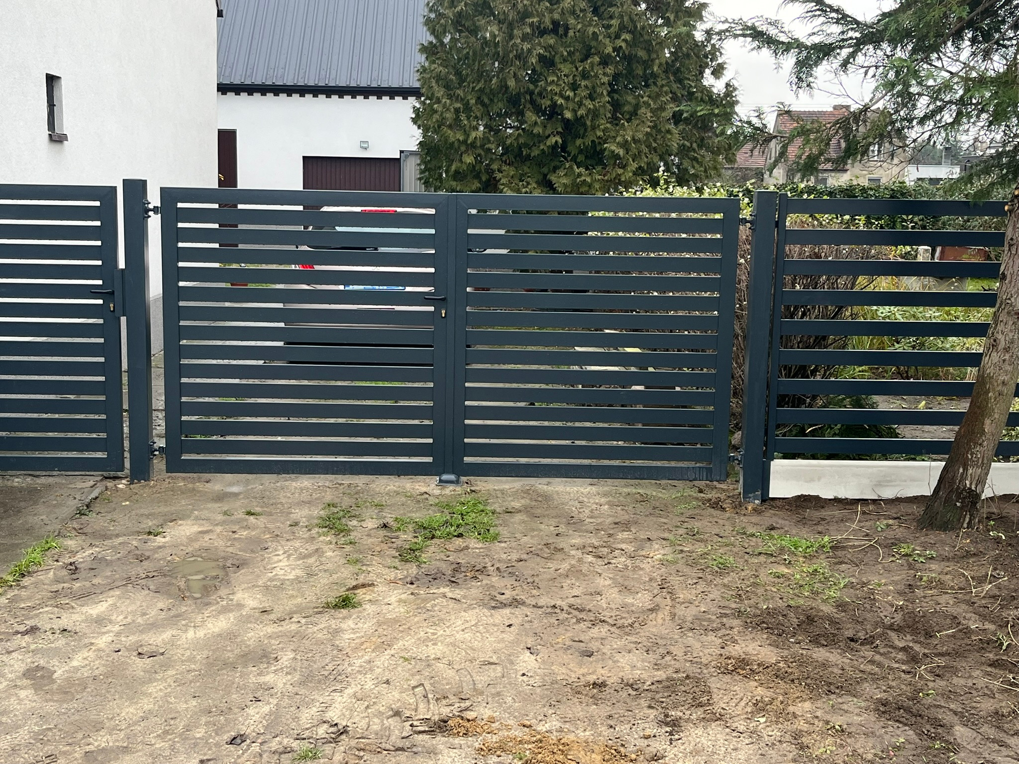 Nowoczesna, pozioma, metalowa brama wjazdowa i ogrodzenie w kolorze grafitowym, z widoczną kłódką, na nierównym, błotnistym podłożu przed domem.