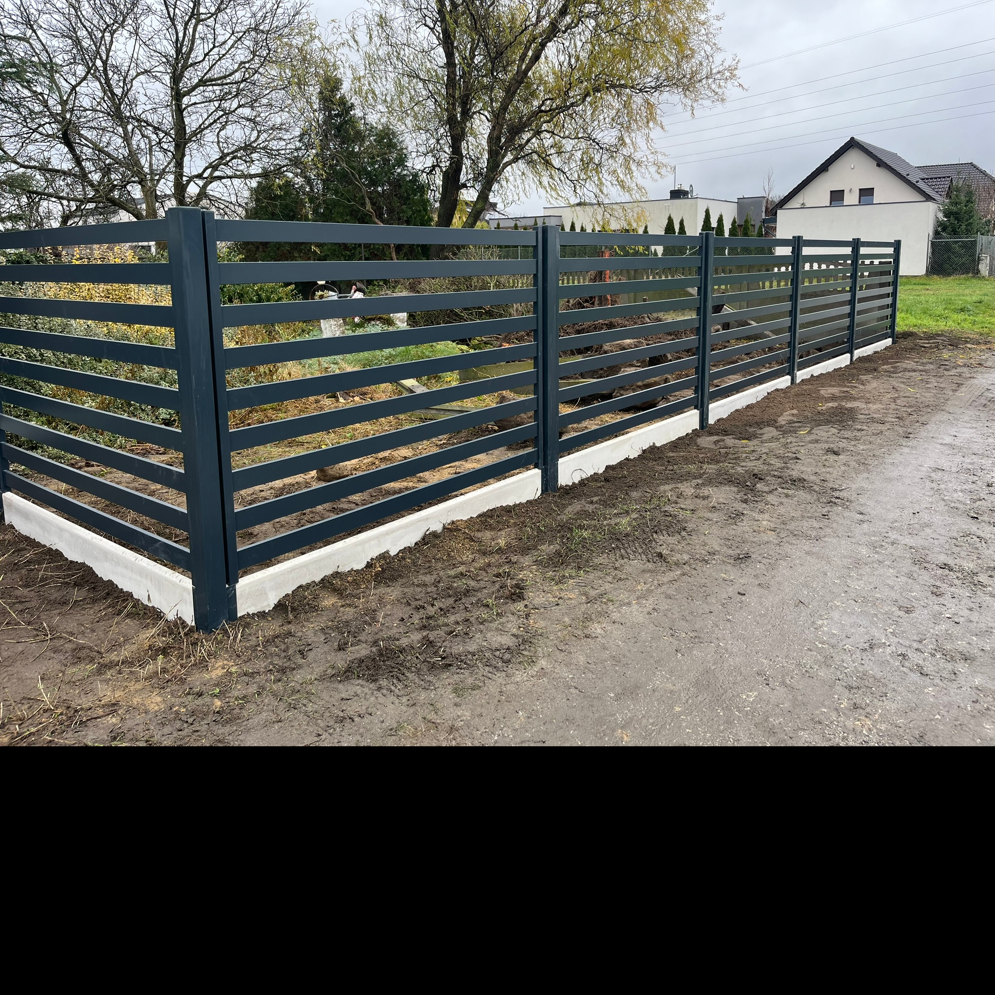 Nowoczesne, poziome ogrodzenie posesji z szarych, metalowych paneli, osadzone na niskiej, betonowej podmurówce, na tle posesji z domem.