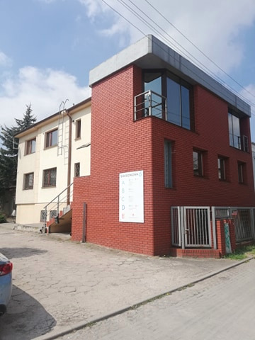 Dwupiętrowy budynek biurowy z ceglaną elewacją i betonowym dachem, obok starszego budynku z zewnętrznymi schodami, tablica informacyjna na ścianie, częściowo widoczny samochód po lewej.