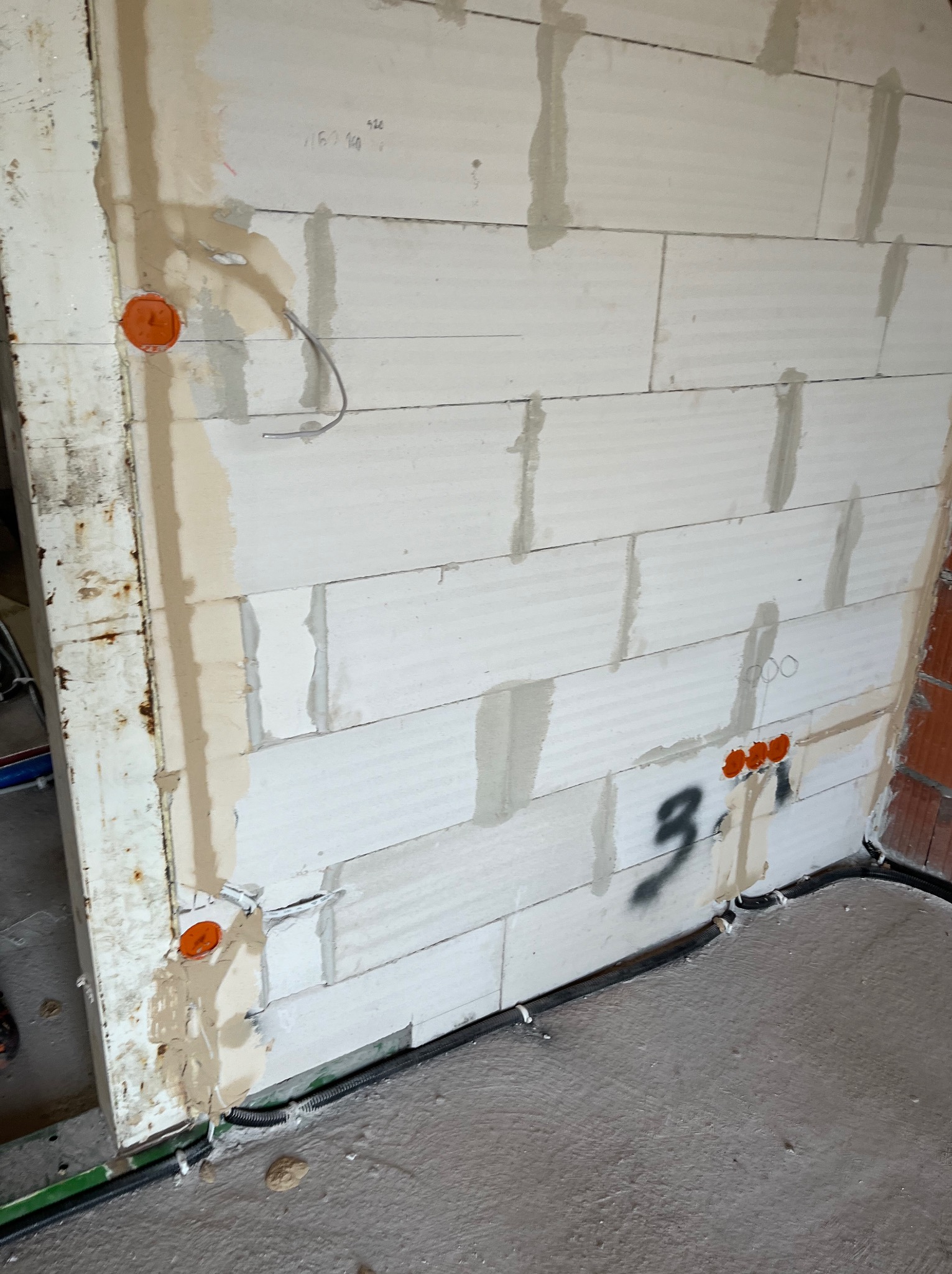 Ściana z betonu komórkowego z widocznymi puszkami instalacyjnymi w kolorze pomarańczowym, przewodami elektrycznymi oraz czarną rurą karbowaną przy podłodze, na ścianie widoczny napis '3.1' wykonany...