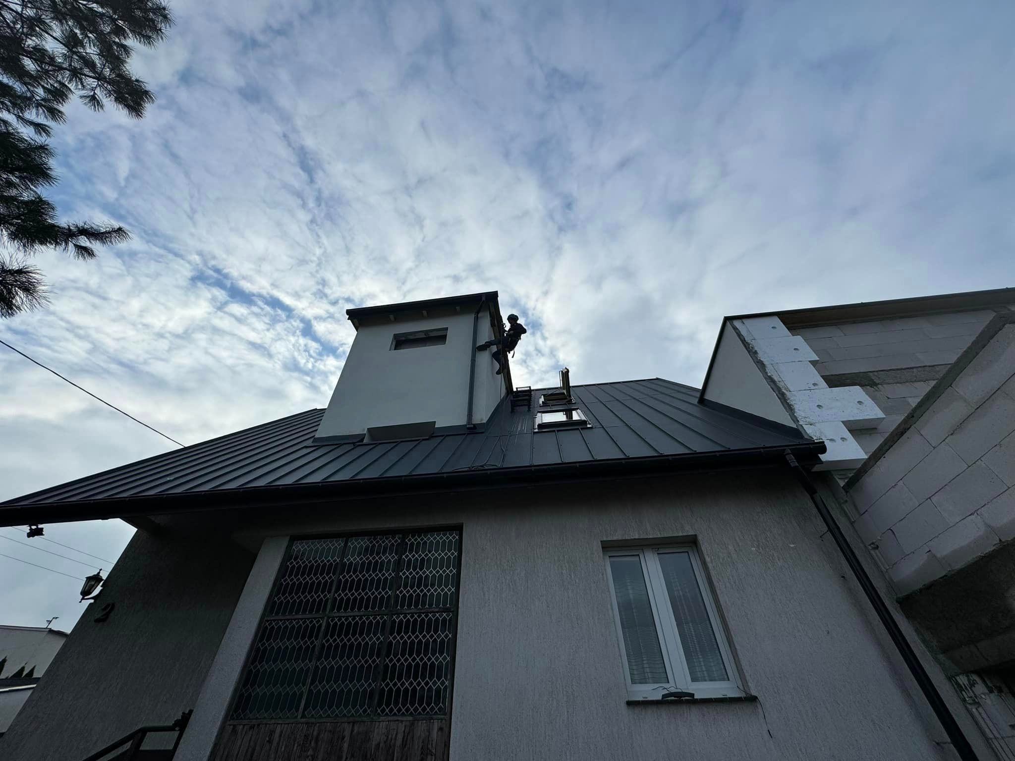 Pracownik zabezpieczony linami, wykonuje prace na wysokości na dachu budynku z blachy trapezowej, widoczny komin i fragment elewacji w trakcie ocieplania.