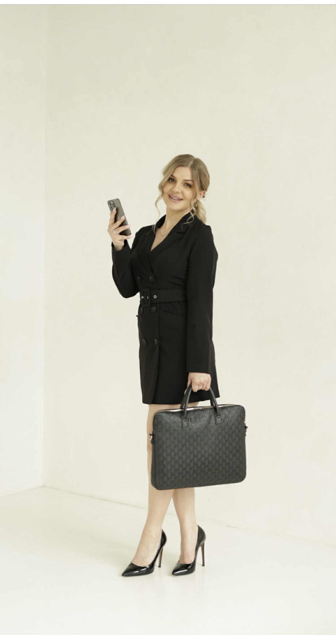 Uśmiechnięta kobieta w czarnej sukience typu marynarka, trzymająca telefon i torbę na laptopa, pozuje na jasnym tle. Widoczne czarne szpilki.