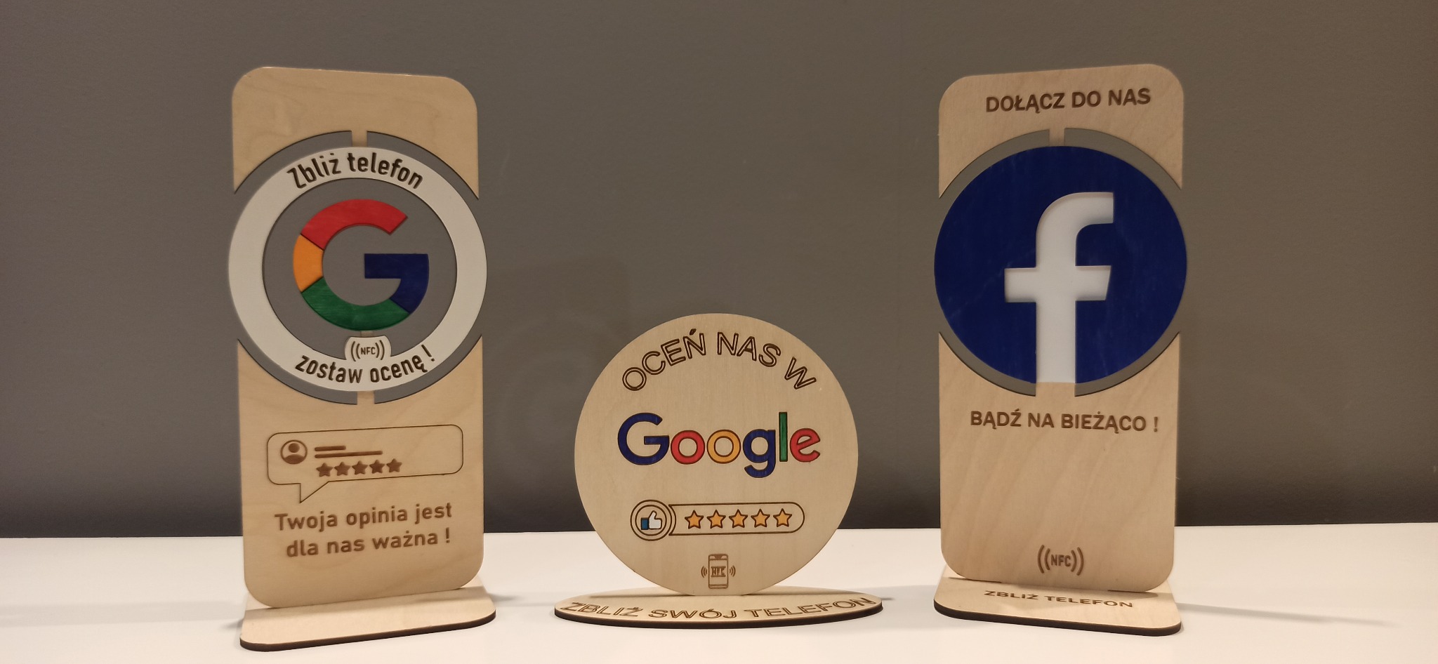 Drewniane standy z logo Google i Facebook z NFC do zbierania opinii. Napisy: 'Zbliż telefon, zostaw ocenę', 'Dołącz do nas, bądź na bieżąco'.