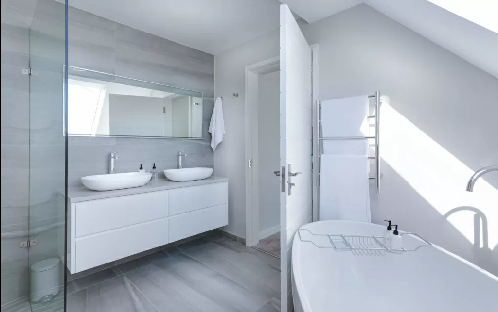 Jasna, minimalistyczna łazienka z dwoma umywalkami na blacie, wanną wolnostojącą i szklaną kabiną prysznicową, utrzymana w odcieniach szarości i bieli, z otwartymi drzwiami i promieniami słońca...